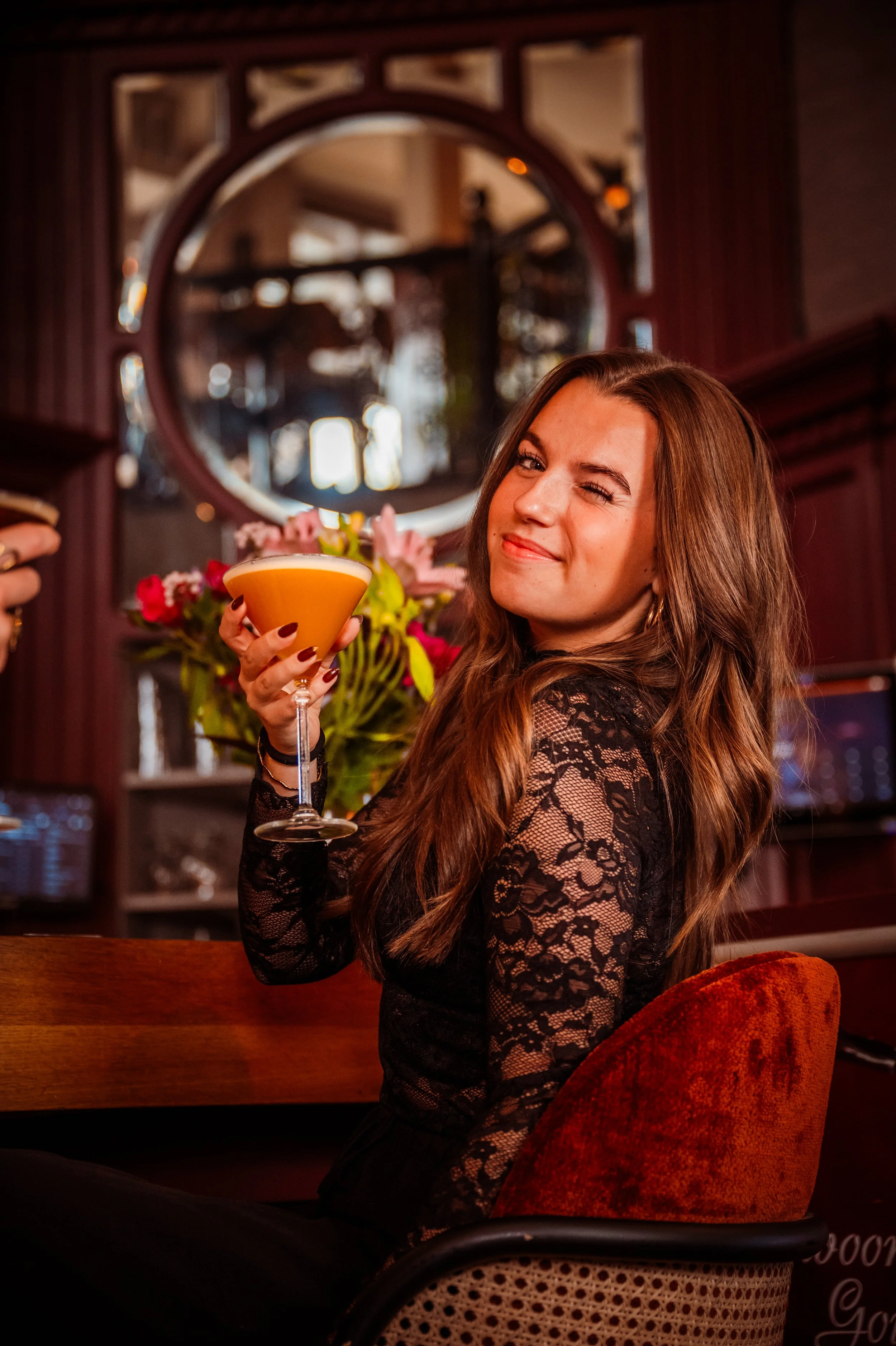 A young woman with long brown hair wearing a black lace top, sitting in a restaurant or bar, winking and holding a cocktail glass with an orange-colored drink. There is a bouquet of flowers and a decorative circular mirror behind her.