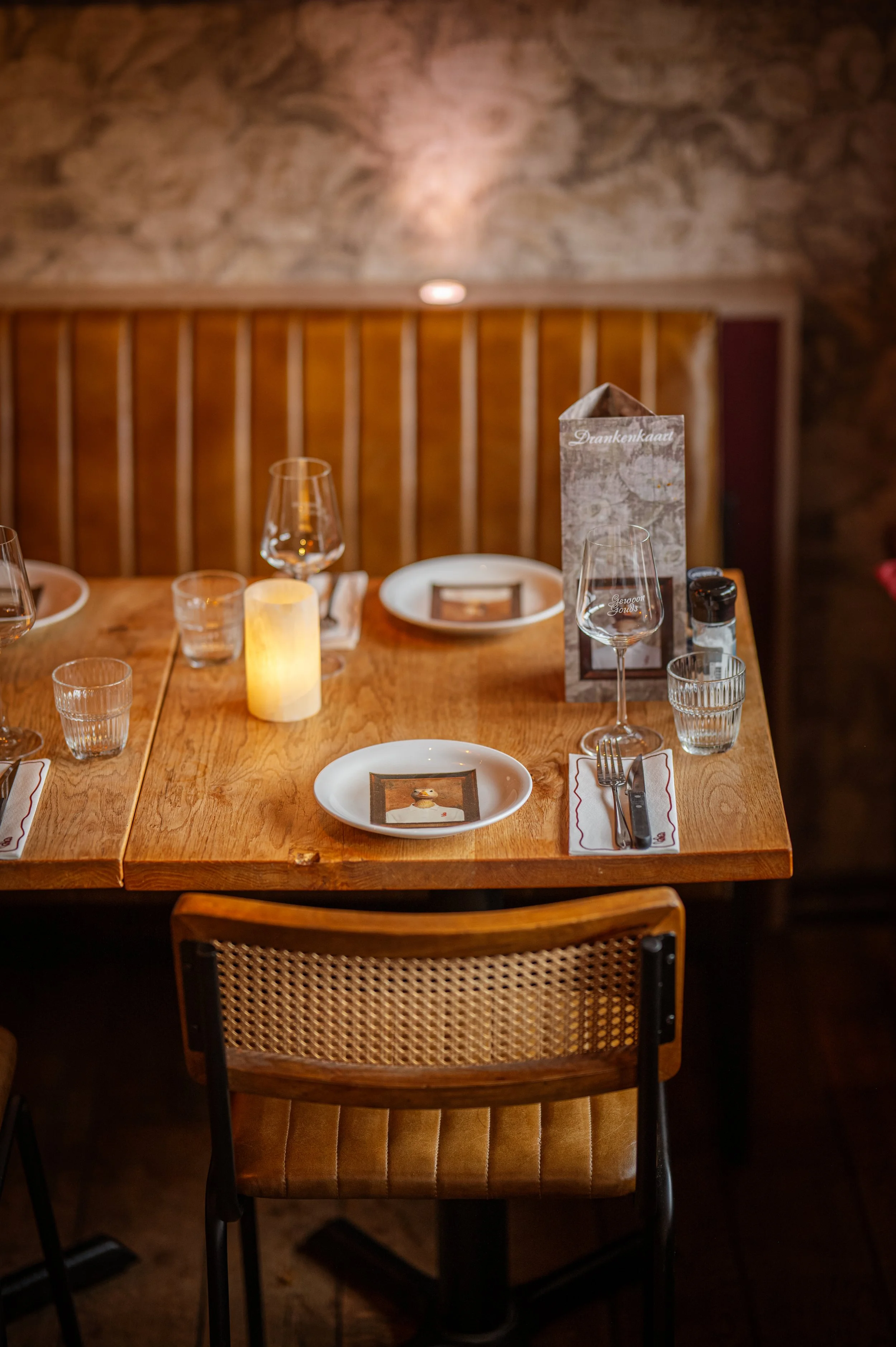 Table set for dining with plates, wine glasses, water glasses, cutlery, a candle, and a menu in a cozy restaurant.