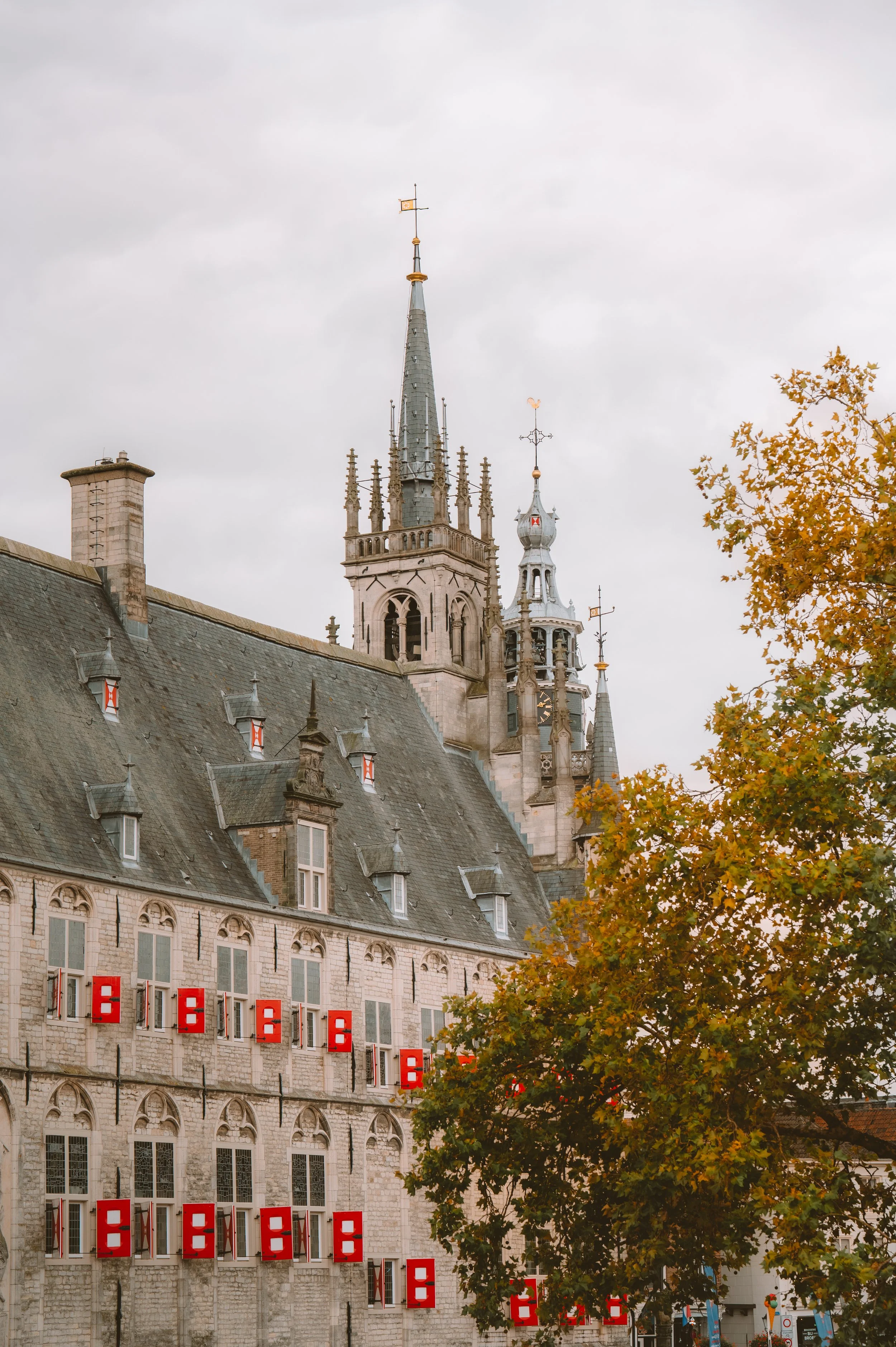 Historic stone building with numerous red window shutters, a tall church spire, and autumn-colored tree in the foreground, under a cloudy sky.
