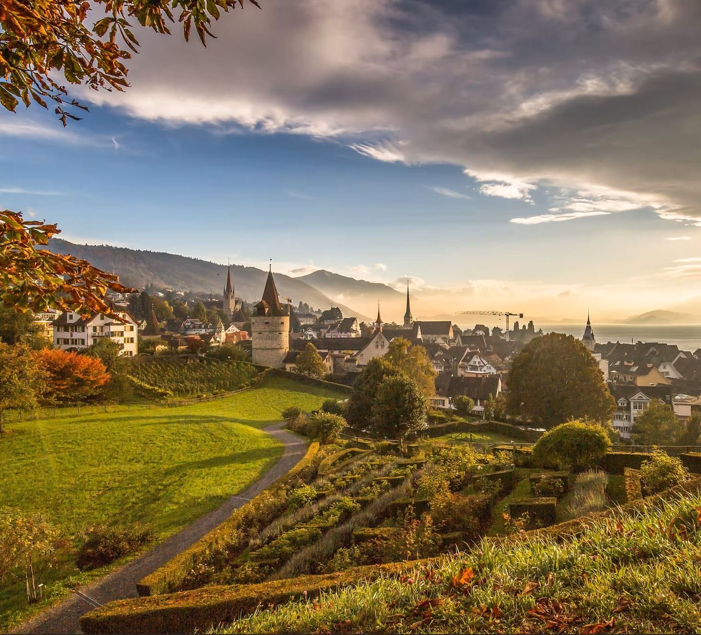Blick auf eine mittelalterliche Stadt mit Kirchtürmen, umgeben von grünem Gelände und Bergen im Hintergrund bei Sonnenuntergang.