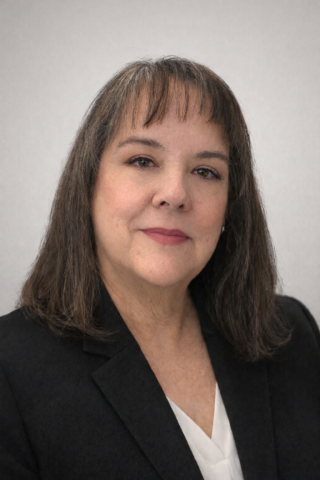 A professional woman with shoulder-length brown hair, wearing a black blazer and white top, smiling with light makeup, posed against a plain light gray background.