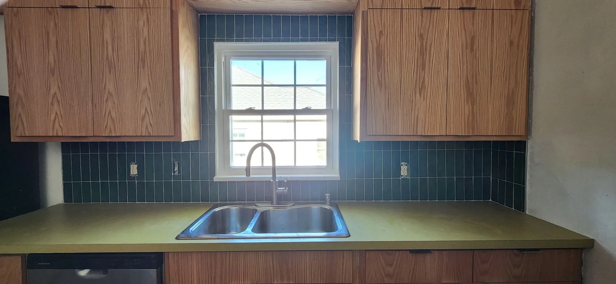 Kitchen with wooden cabinets, green countertop, black tiled backsplash, window above stainless steel double sink, and no appliances or fixtures