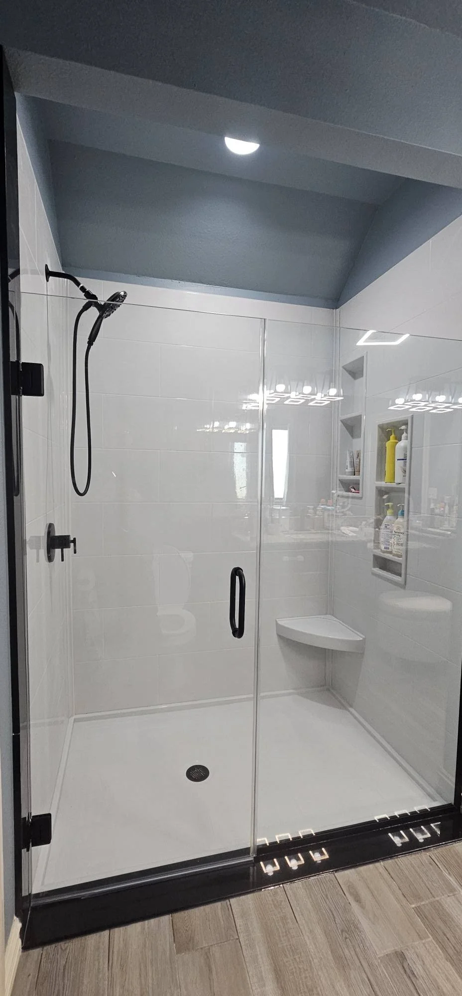 A modern glass-enclosed shower with a black handle, white tiled walls, a black showerhead, a corner shelf with toiletries, and a wooden floor outside the shower.