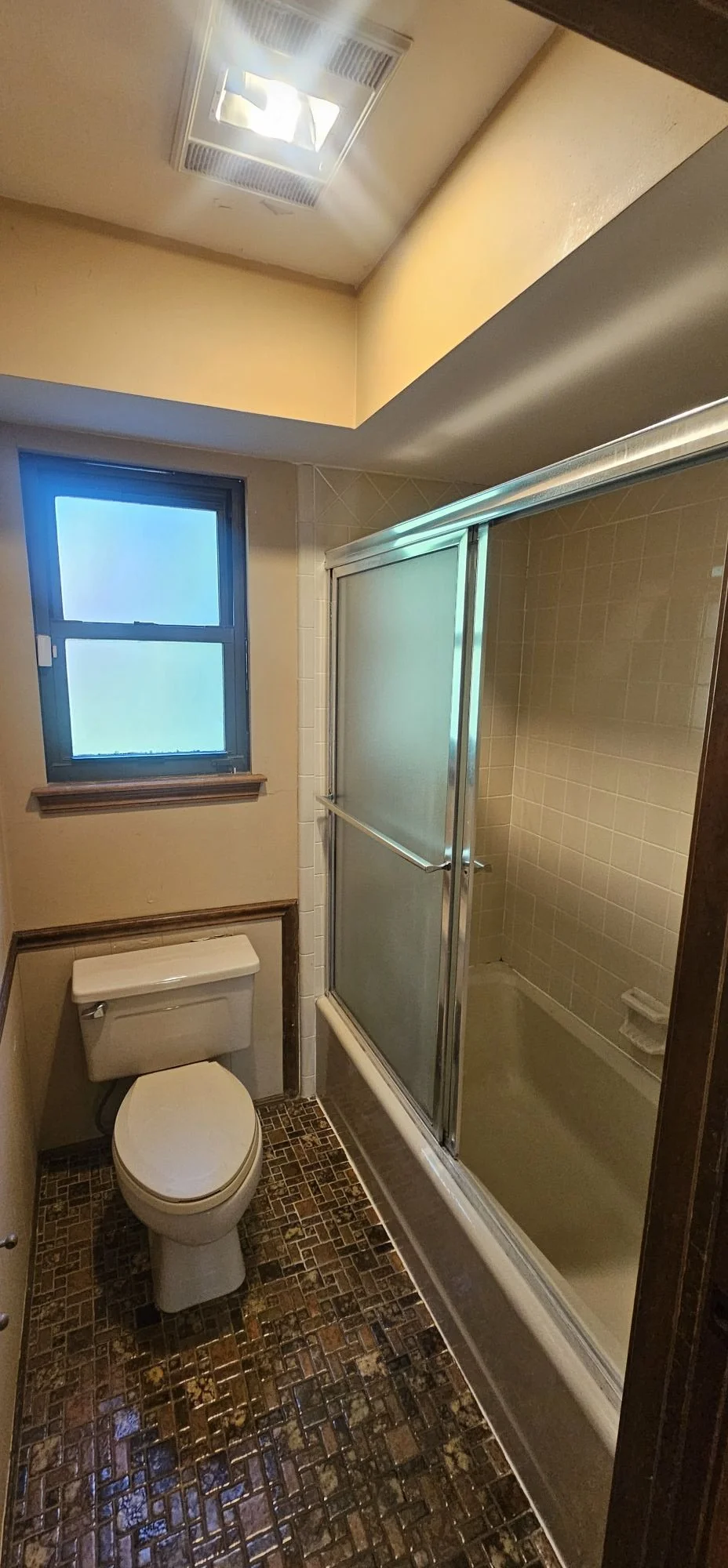 A small bathroom with a toilet, a frosted window, and a shower with a sliding glass door. The floor has a mosaic tile pattern. The walls are painted beige, and there is a ceiling vent.