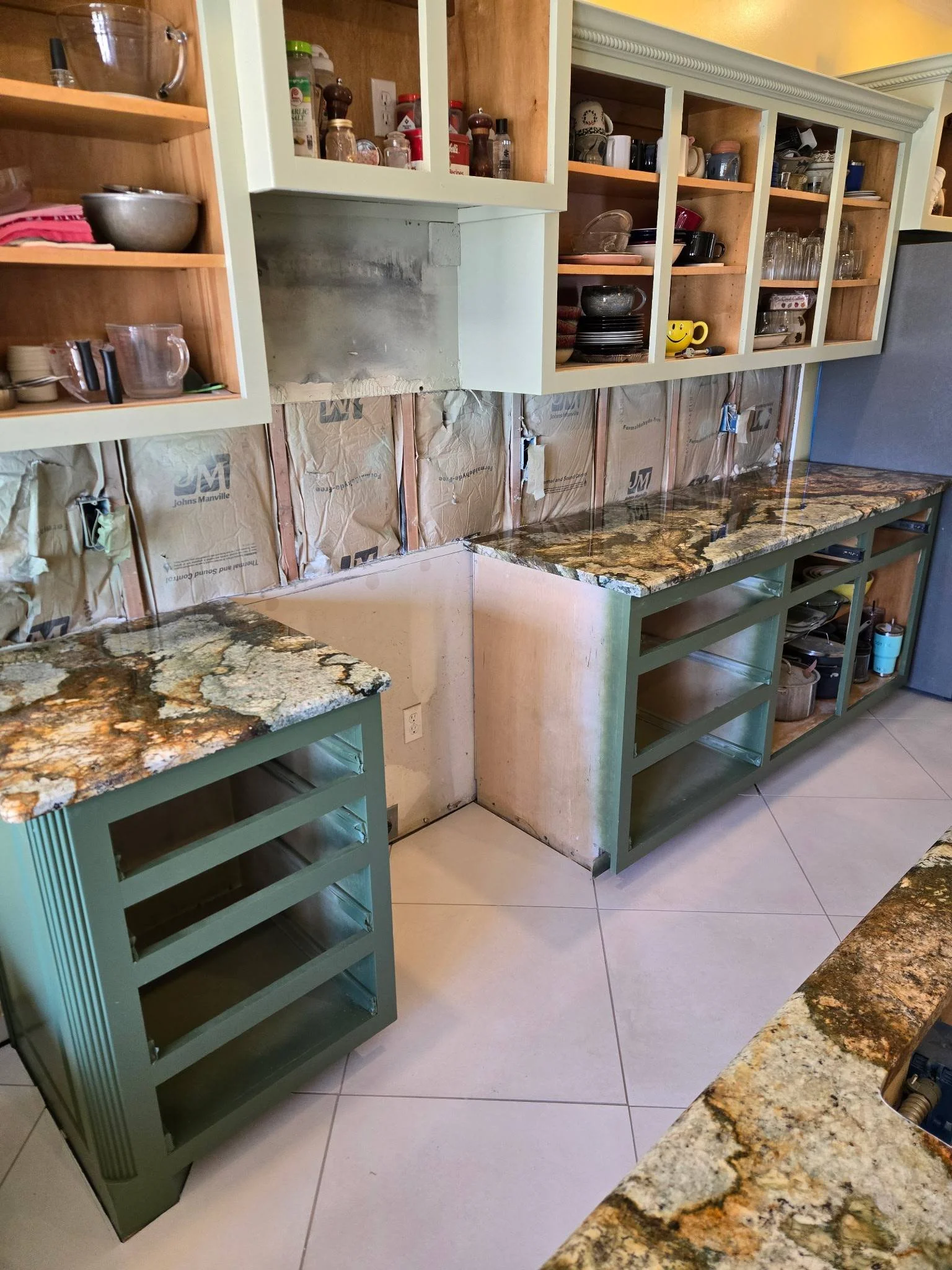 Partial view of a kitchen under renovation with open upper cabinets, marble countertops, and unfinished wall behind cabinets.
