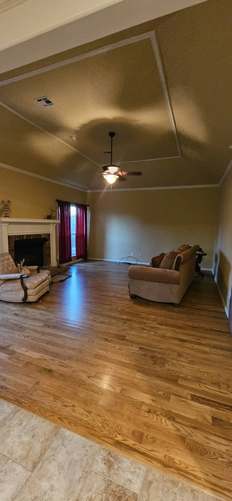 Living room with hardwood floors, a ceiling fan, a fireplace with a mantel, a small sofa with cushions, a chair, and a window with red curtains.