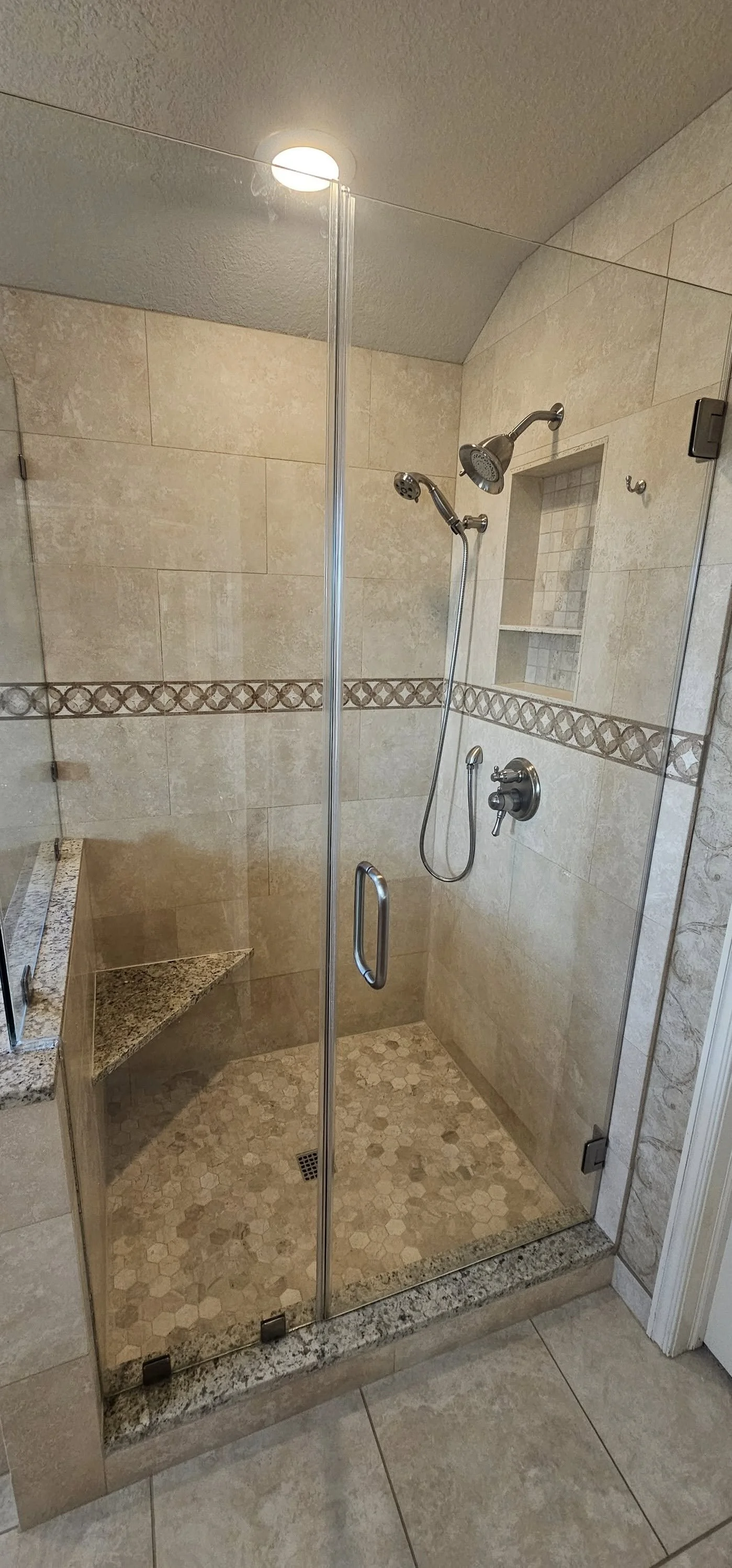 A glass-enclosed shower with two shower heads, a built-in corner bench with a granite top, beige tiled walls with decorative border, and a small niche for toiletries.
