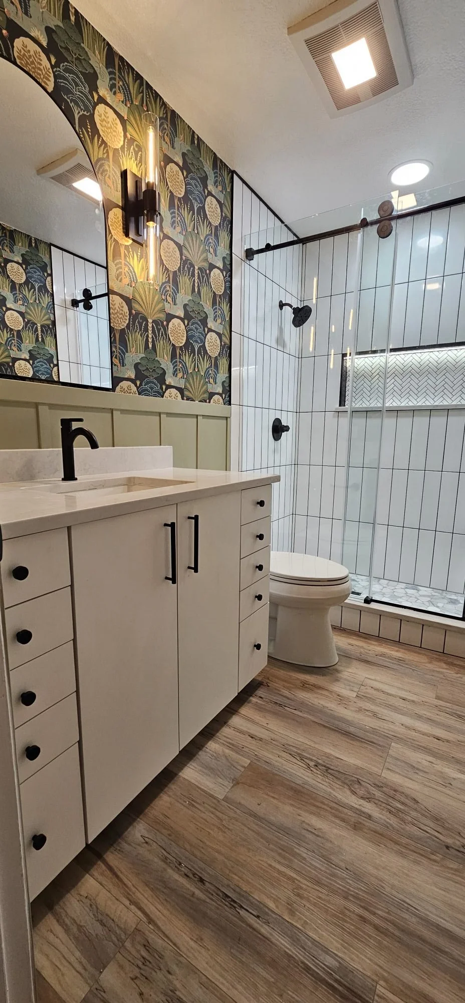 Modern bathroom with wooden flooring, white vanity with black handle fixtures, patterned wallpaper, and a walk-in shower with black fixtures and white tiles.