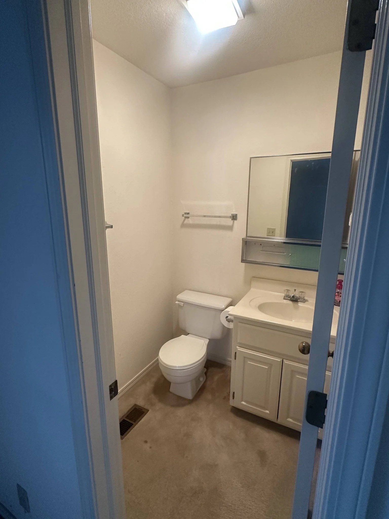 A small bathroom with beige carpet, a white toilet, a white vanity with a sink, a mirror above the sink, a towel bar on the wall, and a ventilation vent on the floor. The bathroom wall is painted in a light color, and there is a ceiling light fixture.