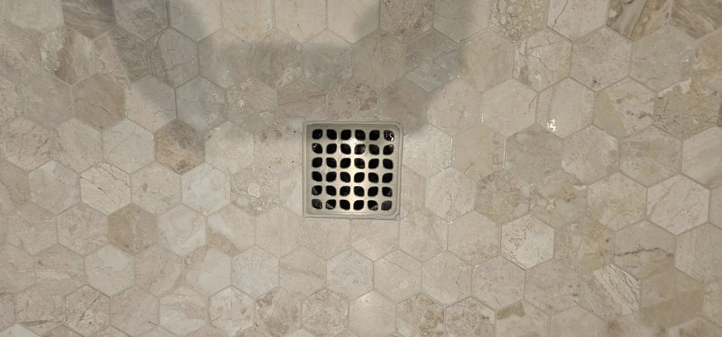 A floor with beige hexagonal tiles and a metal square drain cover in the center.