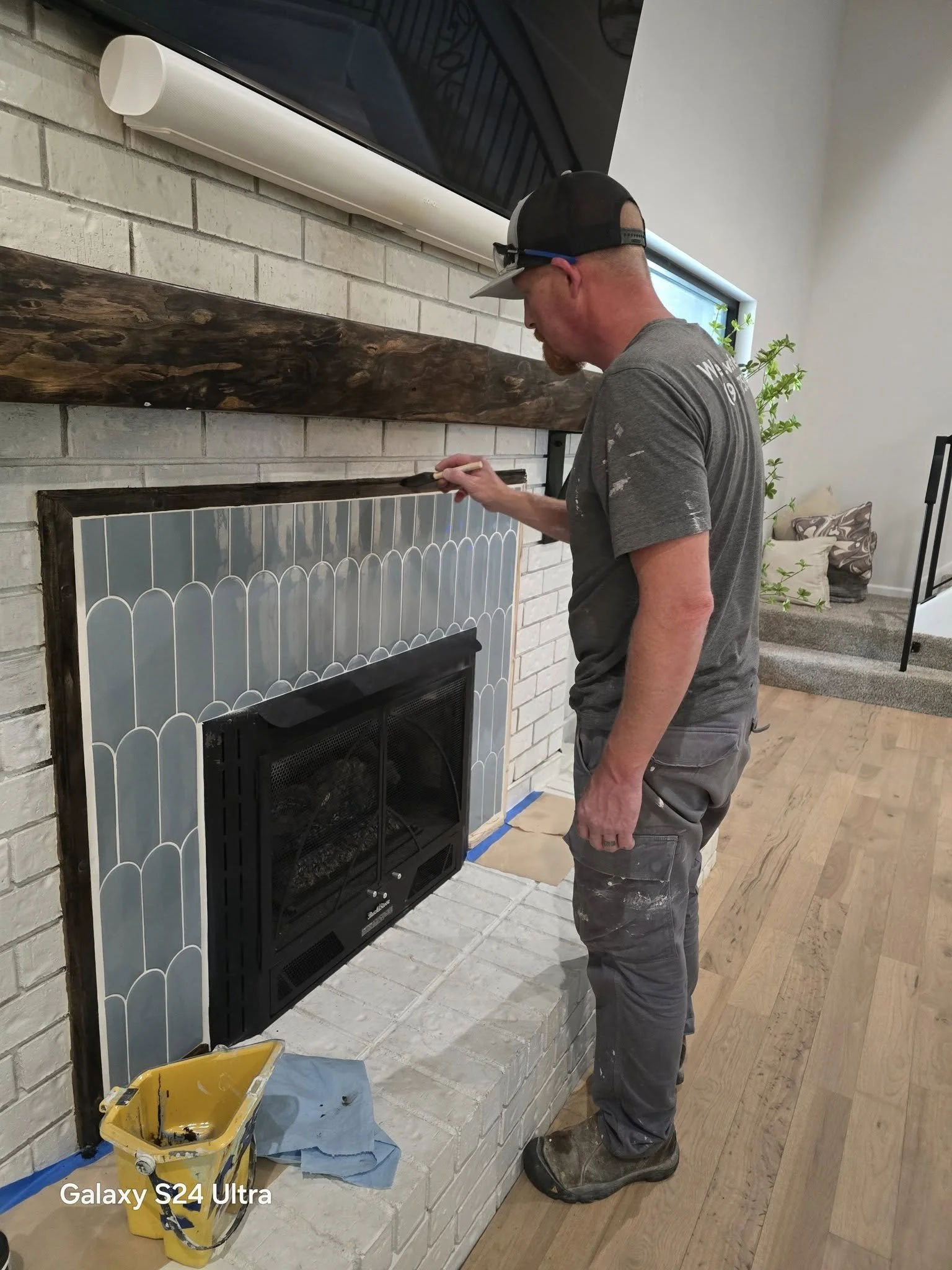 A man wearing work clothes and a baseball cap is painting a brick fireplace with a small paintbrush. The fireplace is finished with white tiles, and the man is adding a dark-colored border around the top. A yellow paint bucket and a blue cloth are on the floor nearby.