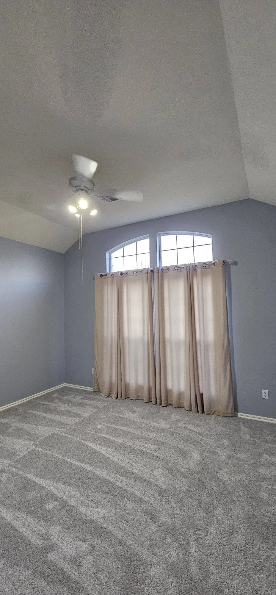 Empty room with light gray walls, beige curtains covering windows, carpeted floor, ceiling fan with light, and a high ceiling.