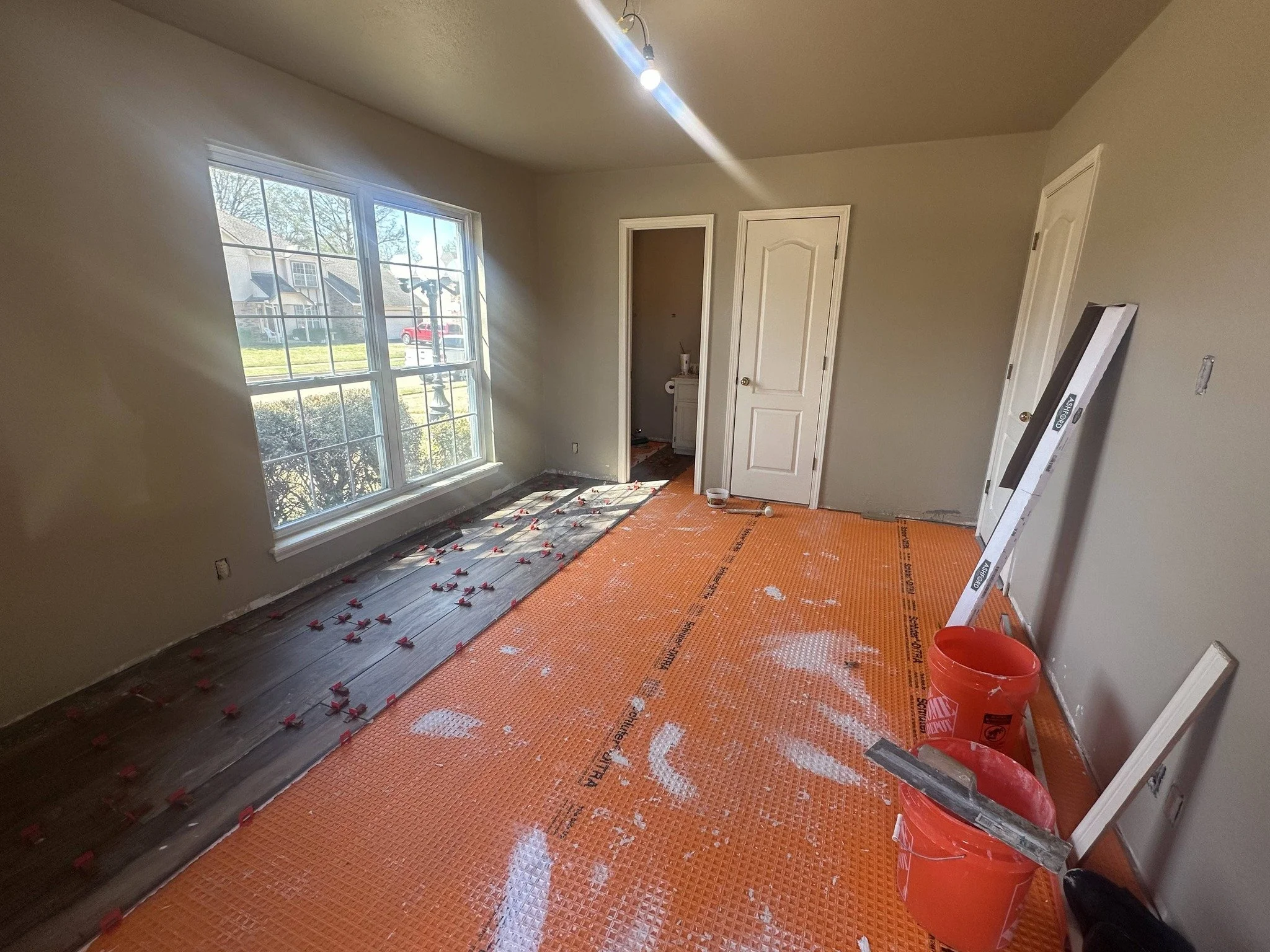 Empty room under renovation with large window, orange construction mat, buckets, and tools.