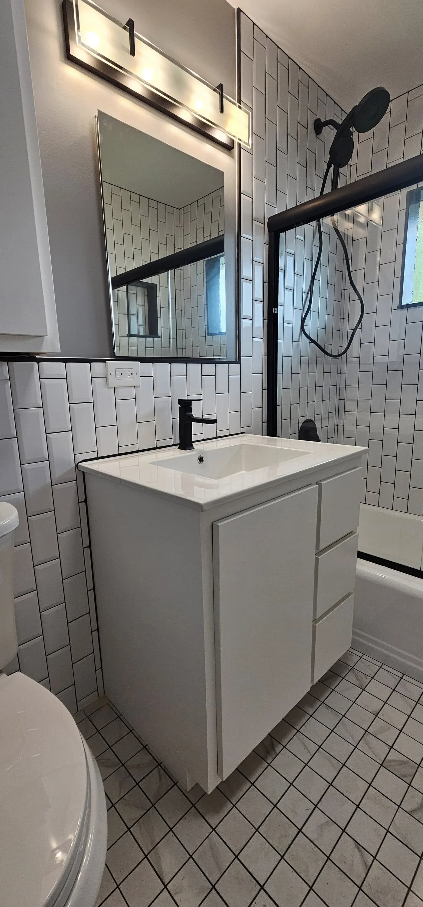 A modern bathroom featuring a white vanity with drawers, a rectangular mirror, tiled walls in a subway pattern, a shower with a black frame, a rainfall showerhead, a small window, and tiled flooring.