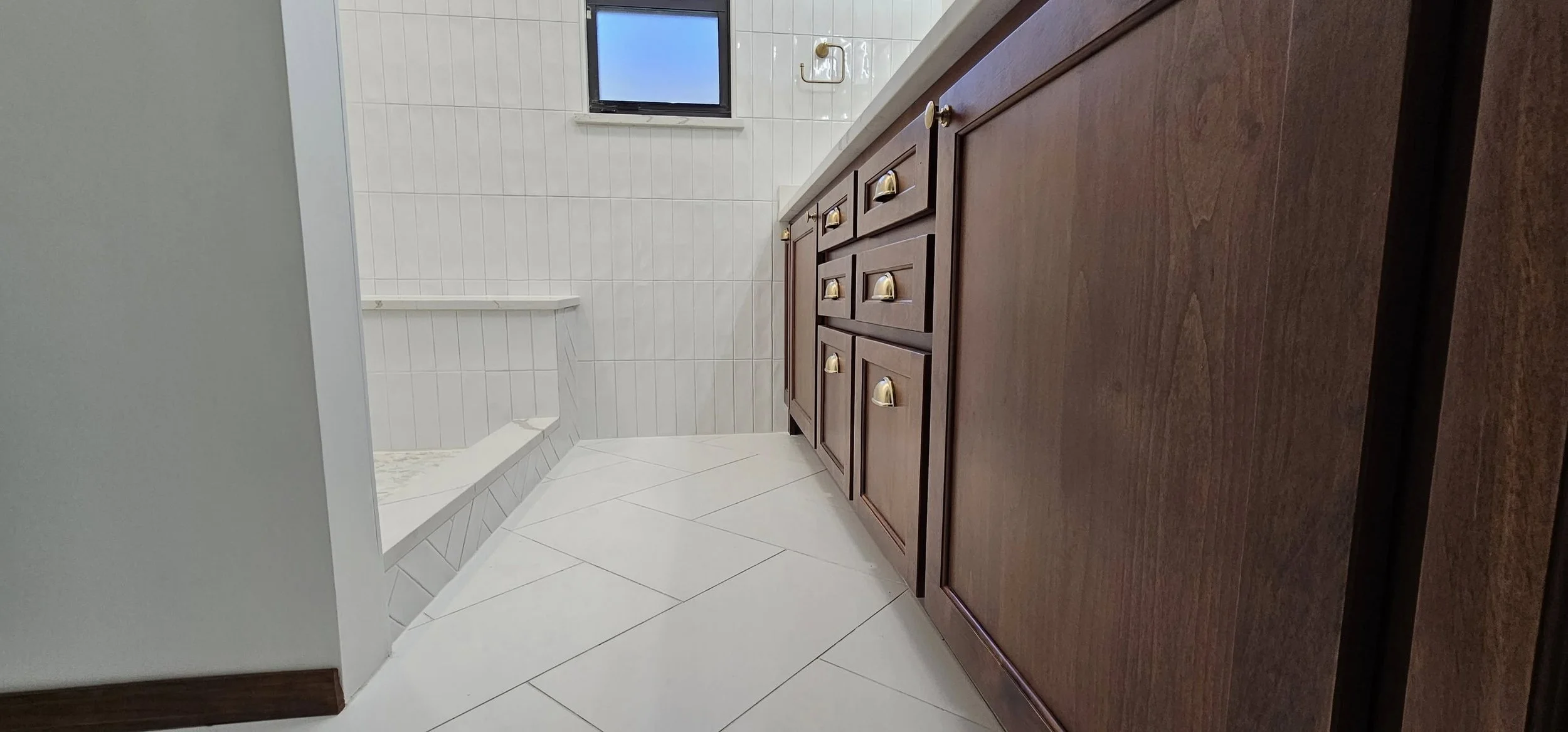 View of a bathroom with a brown wooden vanity with gold handles, white tiled walls, a small window, and a built-in bathtub with white tiling.