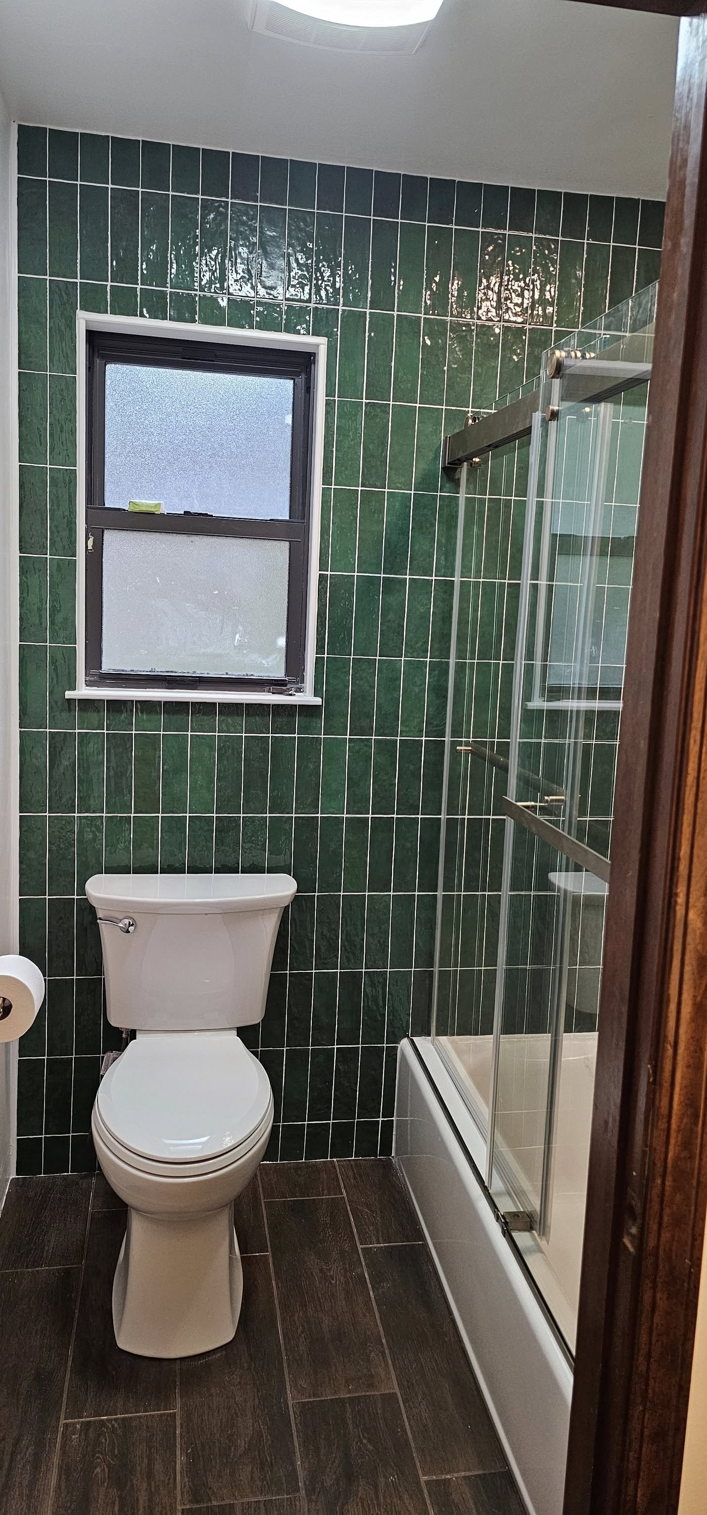 Bathroom with green tiled wall, a frosted window, a toilet, a toilet paper roll, and a glass shower enclosure with sliding door.