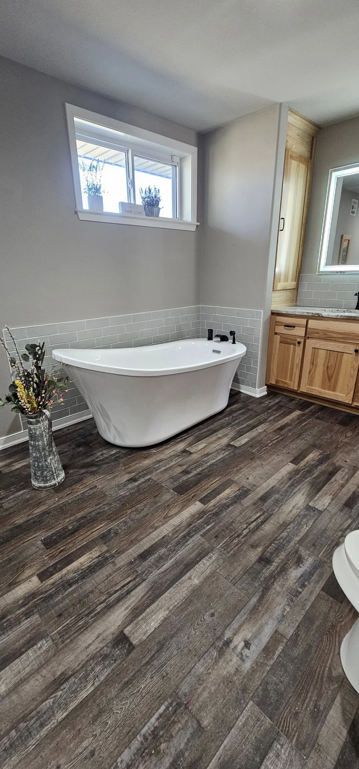 Modern bathroom with a white freestanding bathtub, a window with potted plants, a vase of flowers, wooden cabinets, and wood-look flooring.