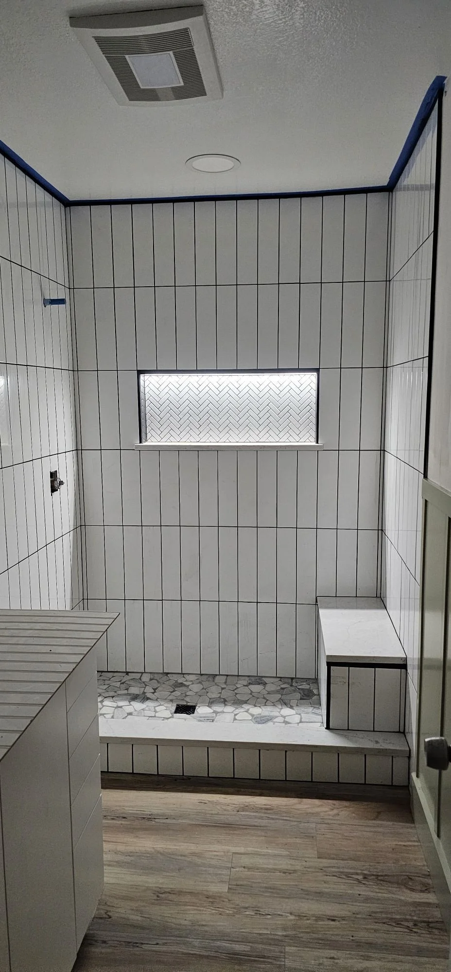 A bathroom with white vertical striped tiles, a small window with a herringbone pattern, and a pebble floor area inside a step down shower space. There is a ventilation fan and a ceiling light.