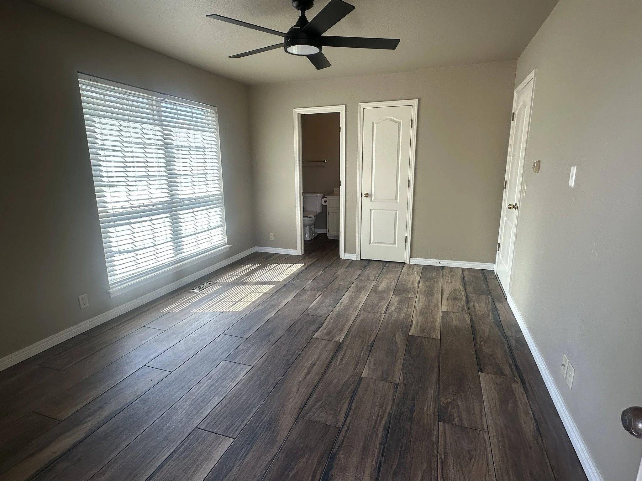 Empty bedroom with hardwood floors, a large window with blinds, and an attached bathroom.