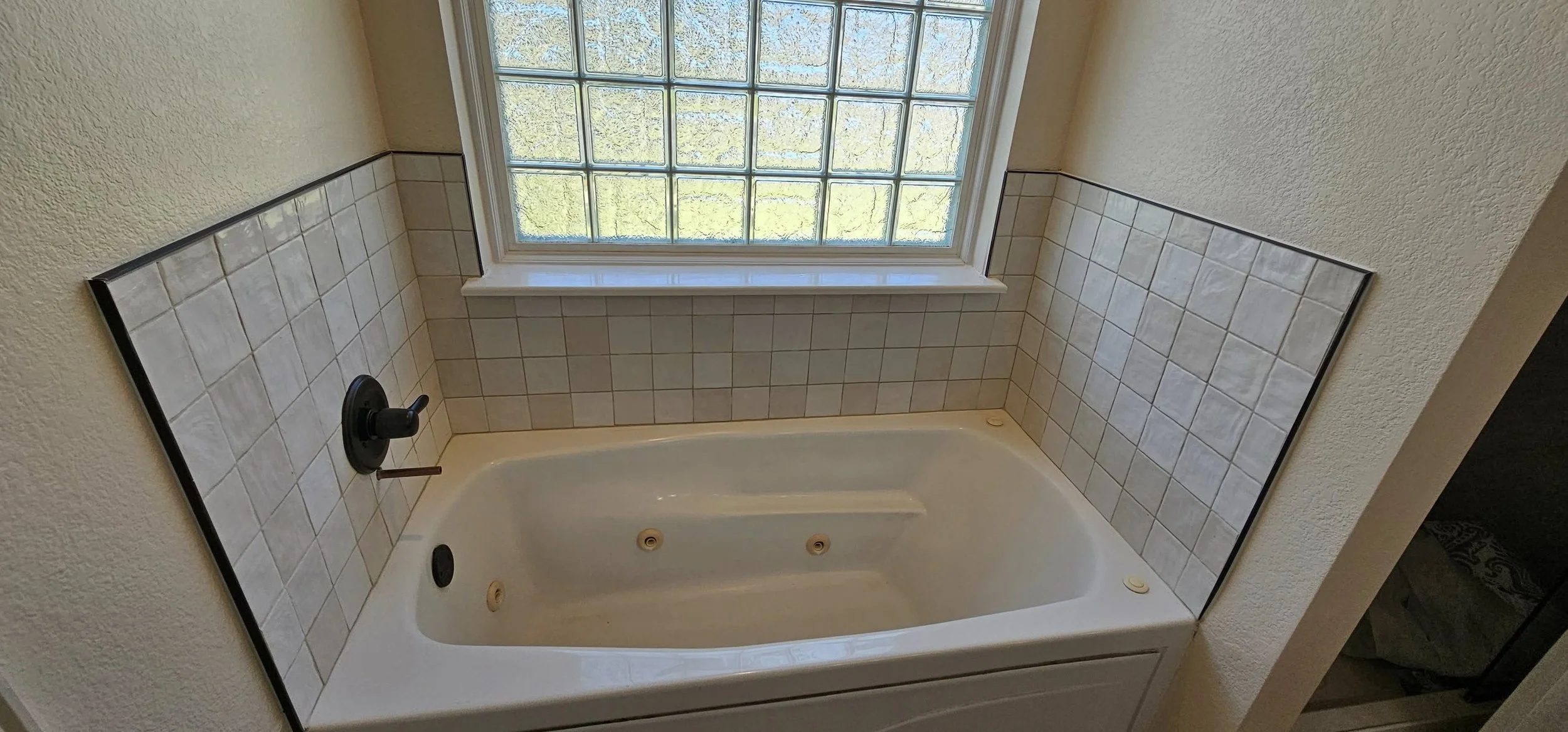 Bathroom with a built-in bathtub, black faucet, tiled wall, and a large window with glass blocks.