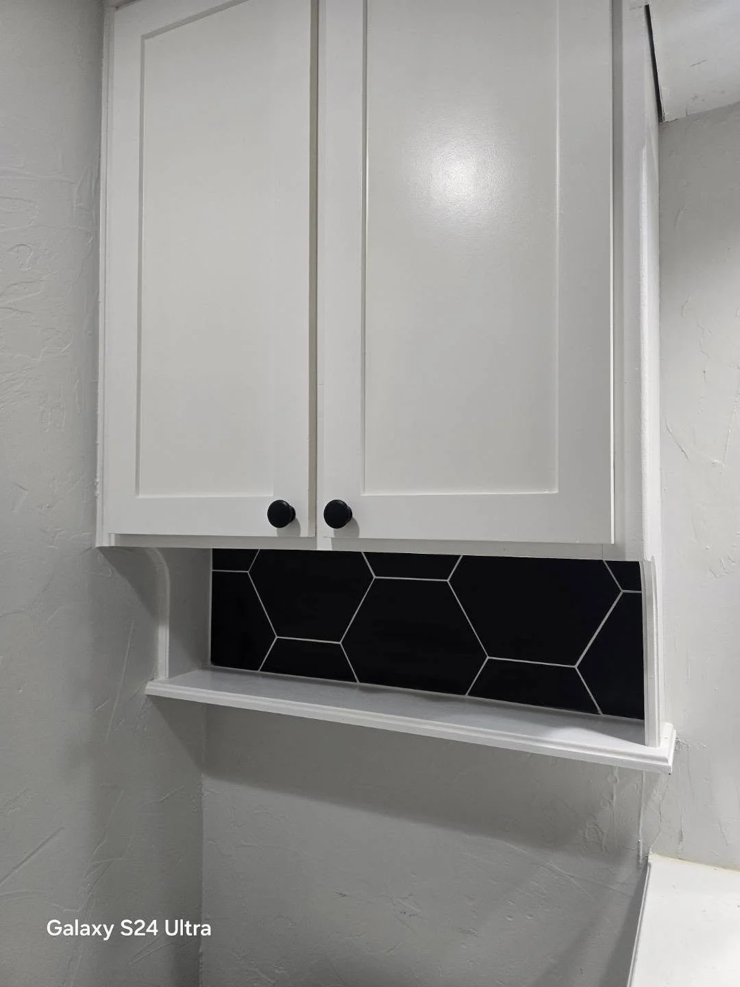 White kitchen cabinet with black knobs, mounted on a wall above a black hexagonal tile backsplash.
