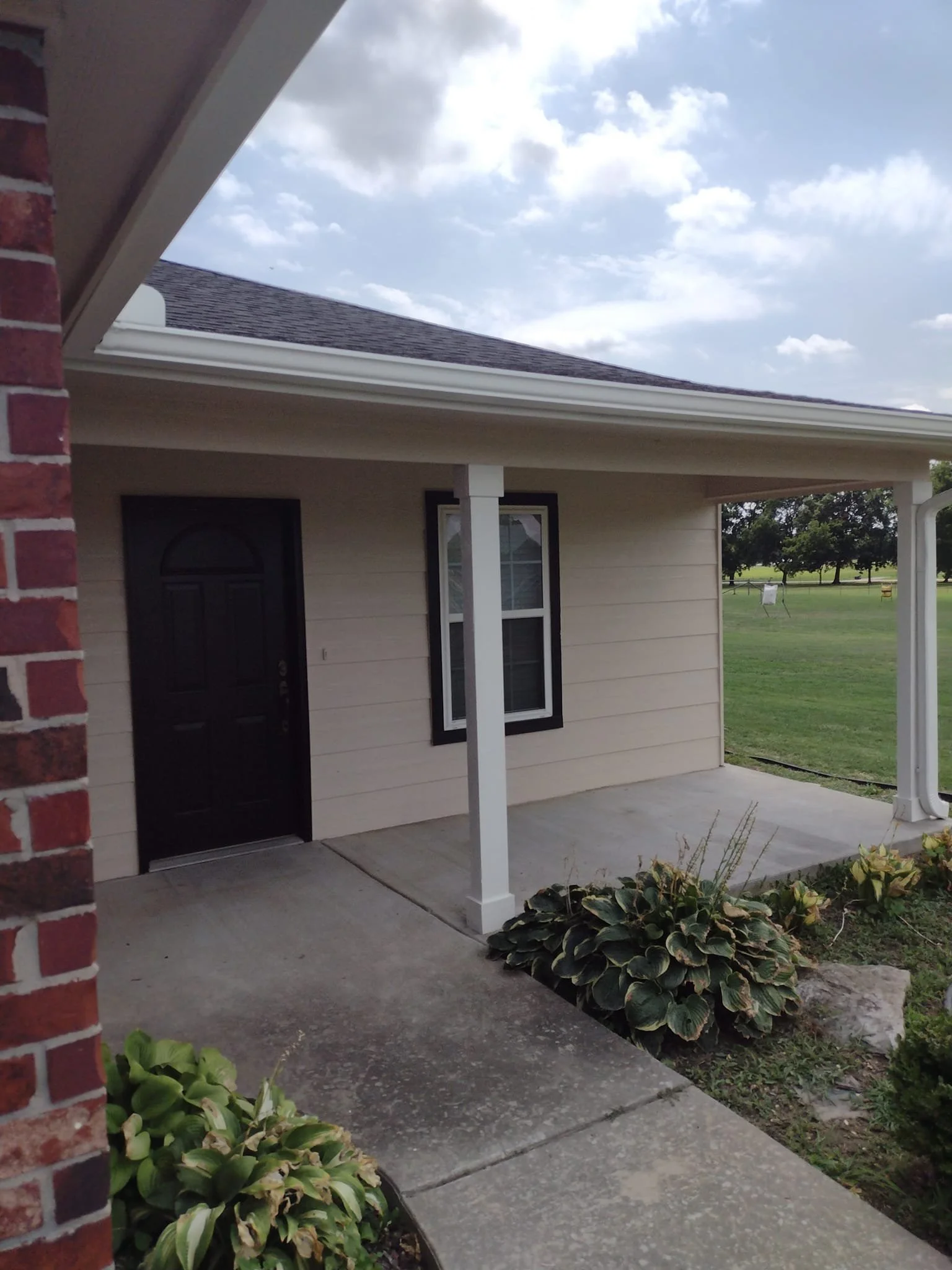 Front porch of a house with a black door, a window with black trim, a white porch post, and a garden bed with green and variegated foliage. The walkway is concrete, and there is a grassy lawn with trees in the background under a partly cloudy sky.