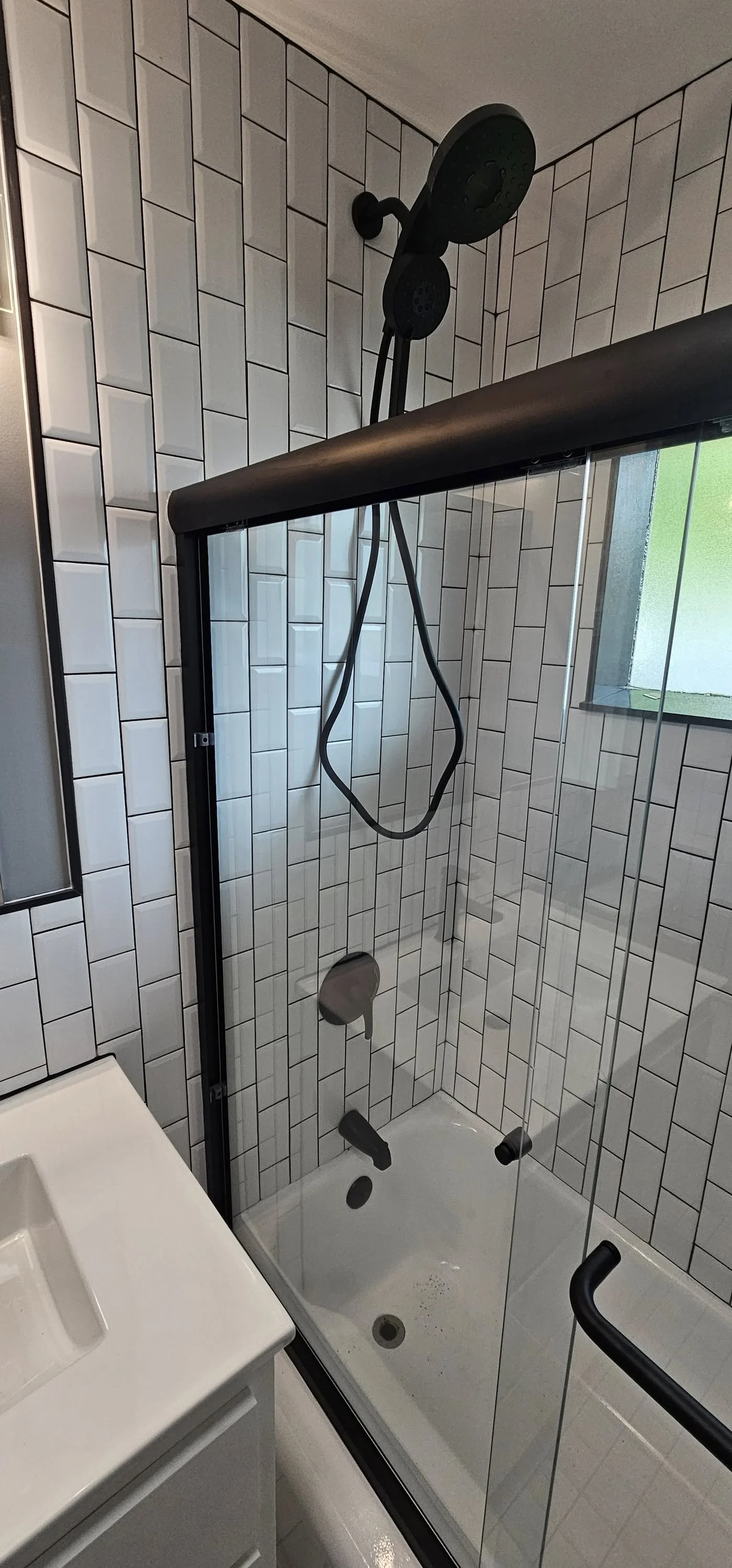 Bathroom shower with black adjustable showerhead, white tiled walls in a brick pattern, a small window with frosted glass, and a glass shower door with a black handle.