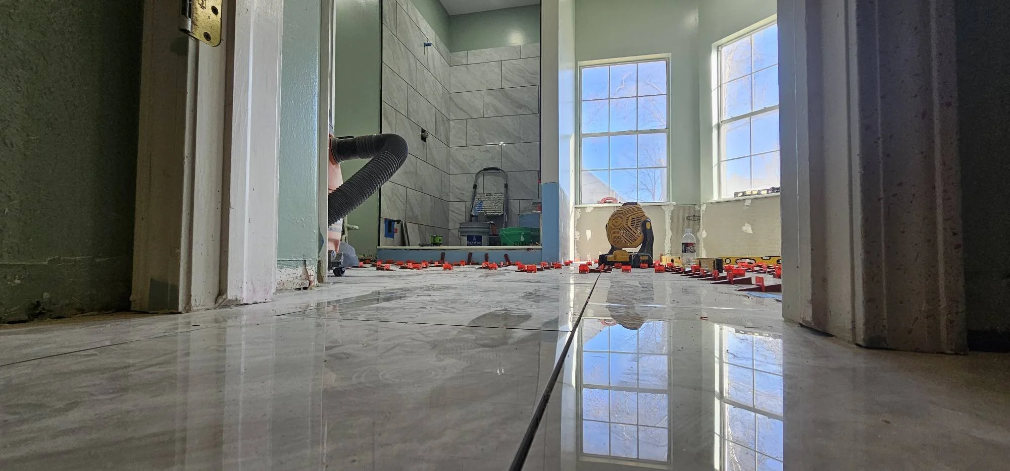 Construction site of a bathroom with tiled floor and wall, showing tools, duct hose, buckets, and large windows allowing natural light.