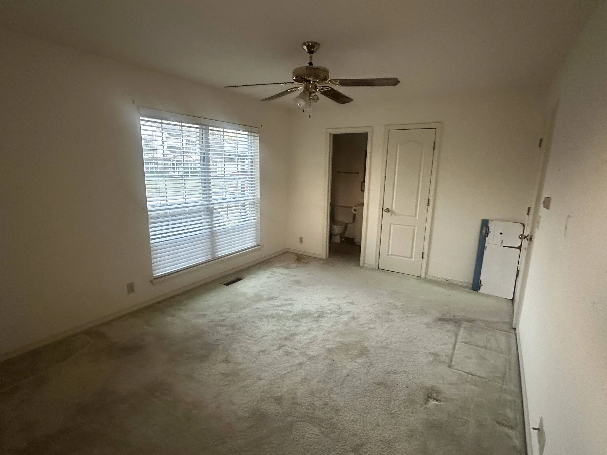 Empty living room with white walls, a large window with blinds, a ceiling fan, beige carpet, and two doors leading to a bathroom and storage closet.