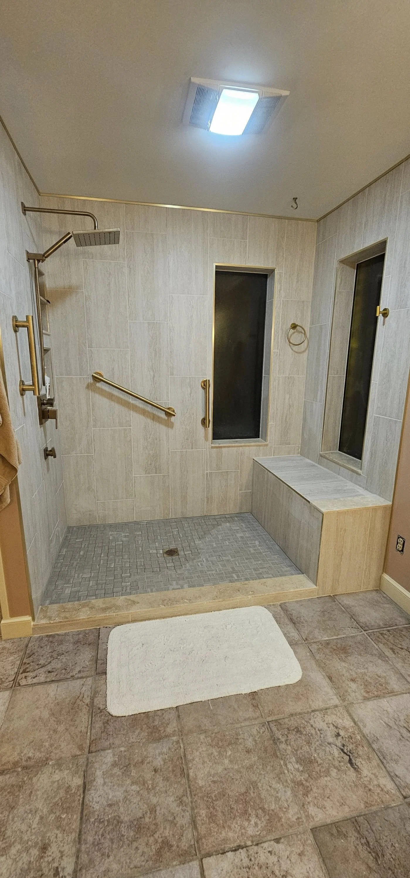 A walk-in shower with beige tiled walls, a small bench, two narrow windows, a ceiling light, and gold-colored grab bars. The shower flooring is gray tile with a drain, and a white bath mat is placed outside the shower on a tiled floor.