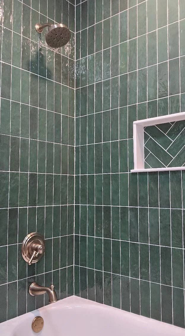 Green tiled shower with a round showerhead, a bronze faucet, and a built-in shelf with a herringbone tile pattern.