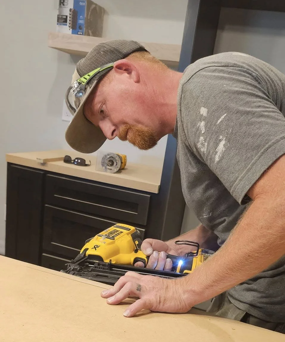 A man with a baseball cap, safety glasses, and a tattoo on his hand is drilling a hole in a wooden workbench with a yellow cordless drill. He appears to be focused on the task.