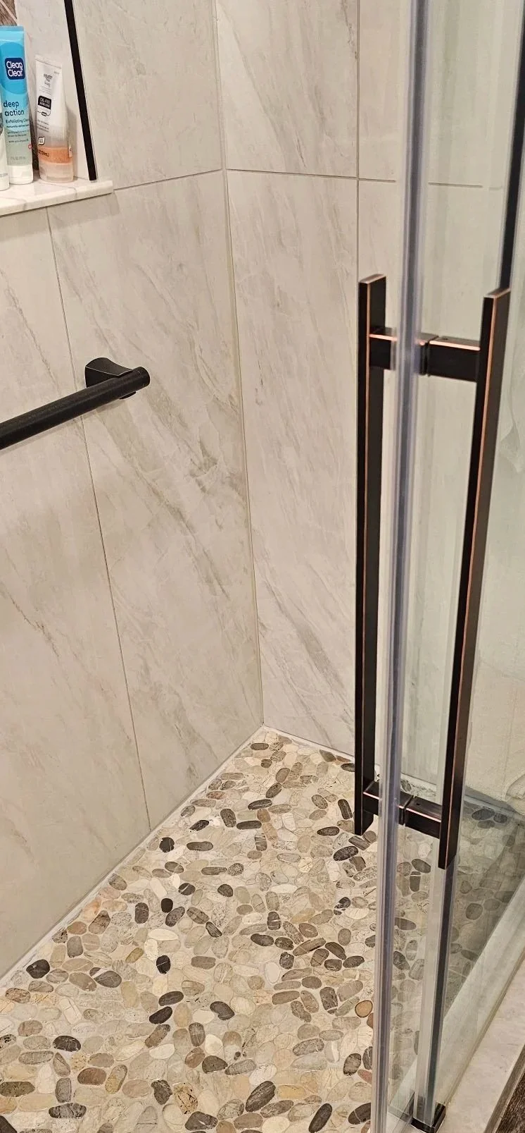 View of a modern shower area with beige marble wall tiles, pebble stone floor, black grab bar, and a glass door with black handles.