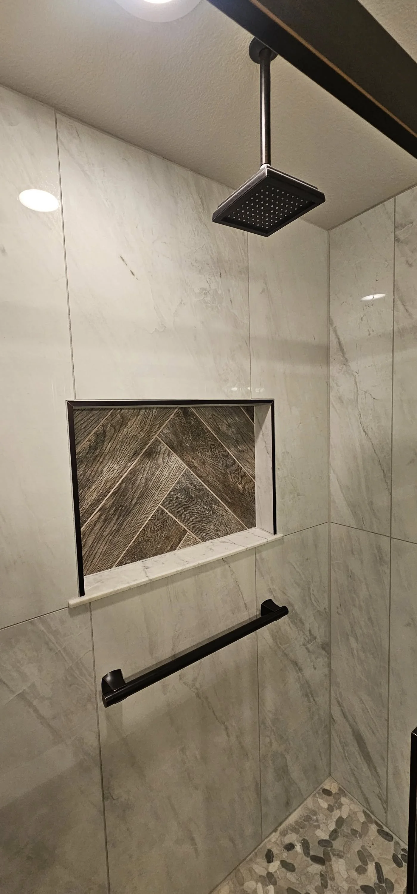 Shower with a rainfall showerhead, a niche with wooden tile design, a black grab bar, ceramic wall tiles, and pebble floor tiles.