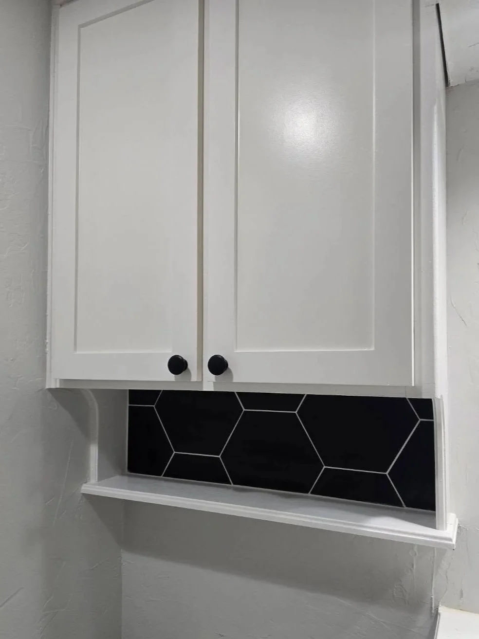 White bathroom cabinet with black knobs above a black hexagon tile backsplash on a light-colored wall.