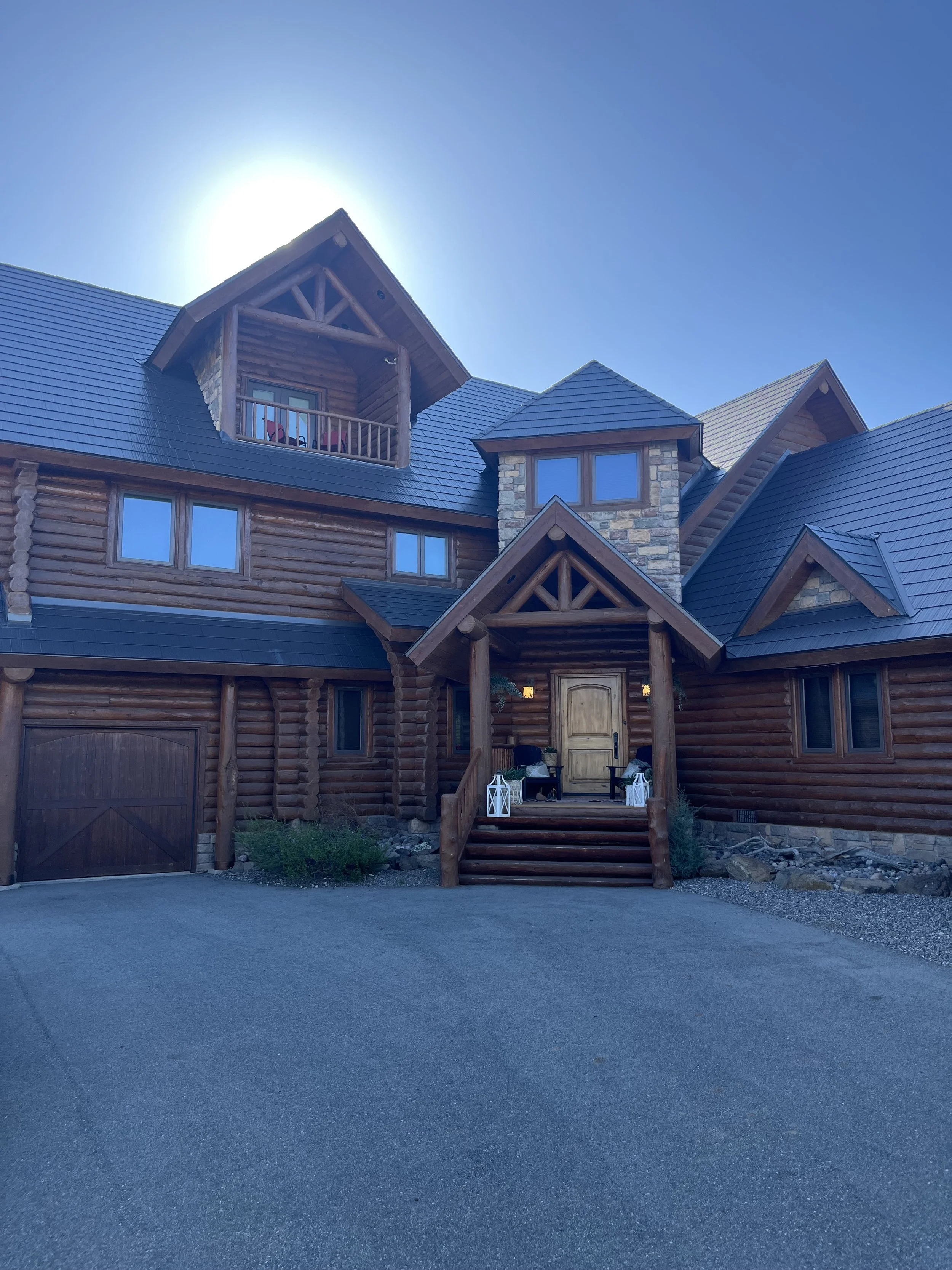 A large wooden house with a stone chimney, multiple windows, and a covered front porch with steps decorated with lanterns, under a clear blue sky with the sun shining behind the roof.