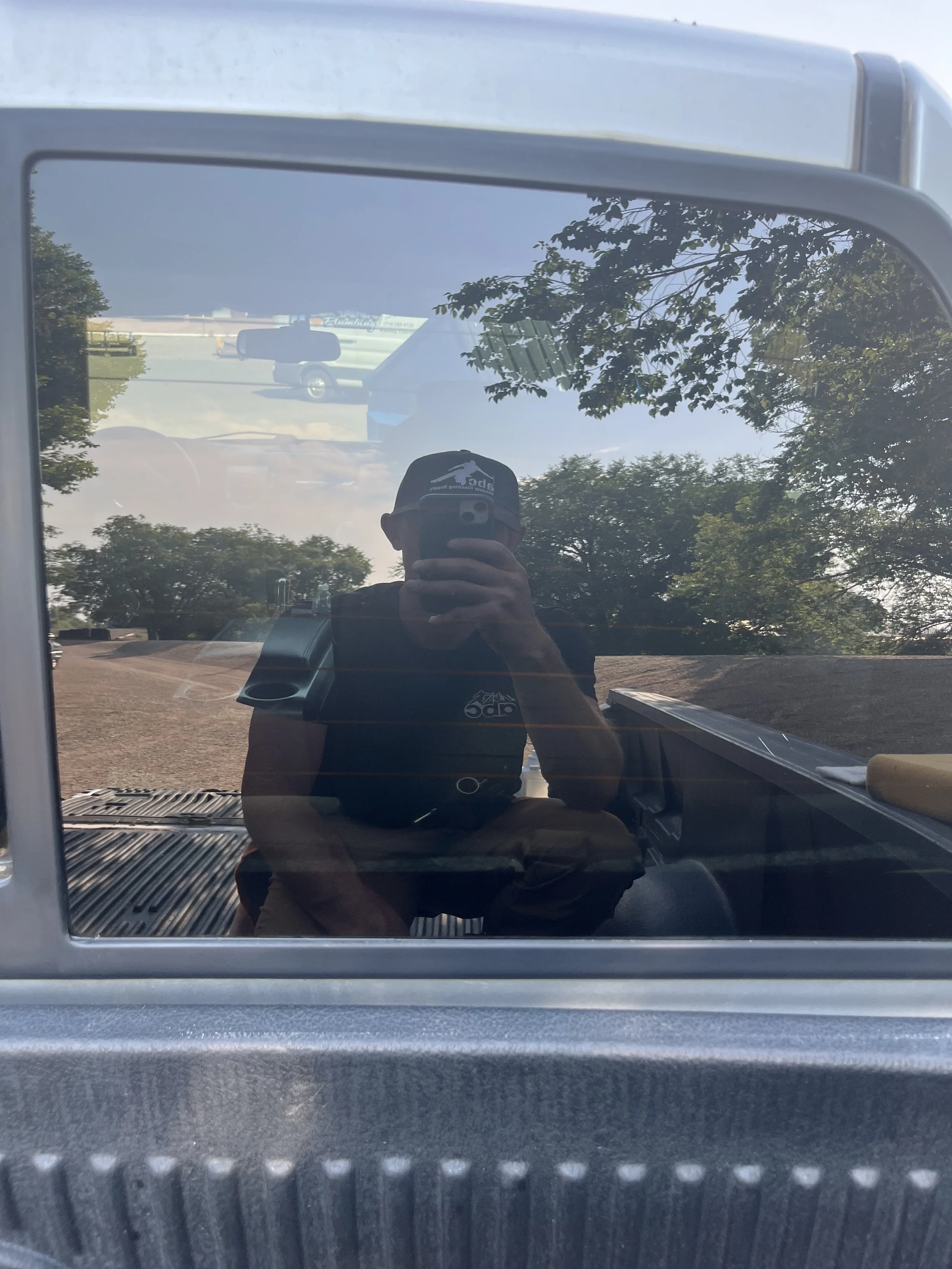 Reflection of a person wearing a black cap and black T-shirt, taking a photo with a smartphone in a mirror, with trees, a blue sky, and vehicles in the background.