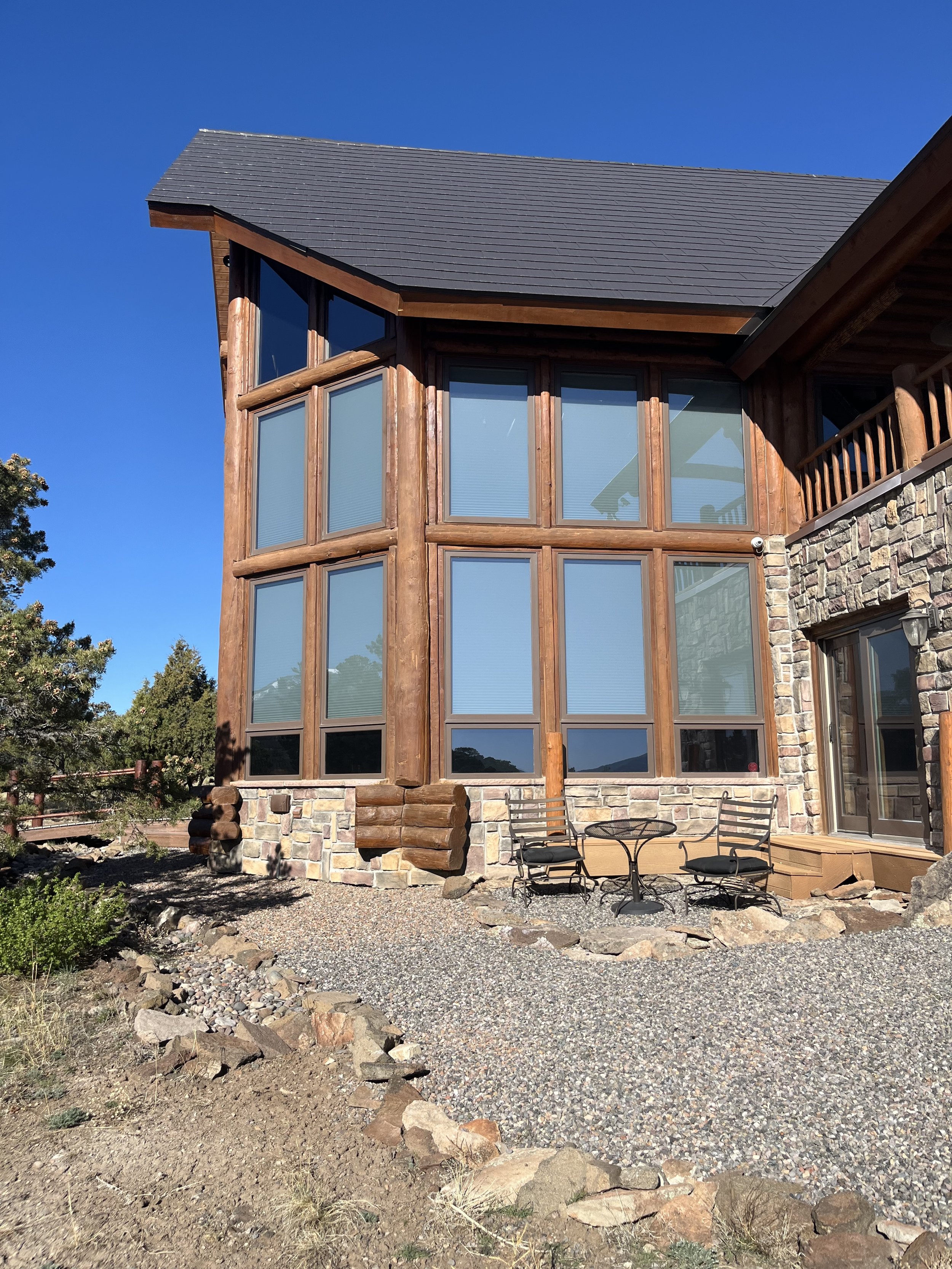 A two-story house with large front windows, a wooden balcony, and a stone and wood facade under a clear blue sky. There are outdoor chairs and a table on a gravel and rocky yard area.
