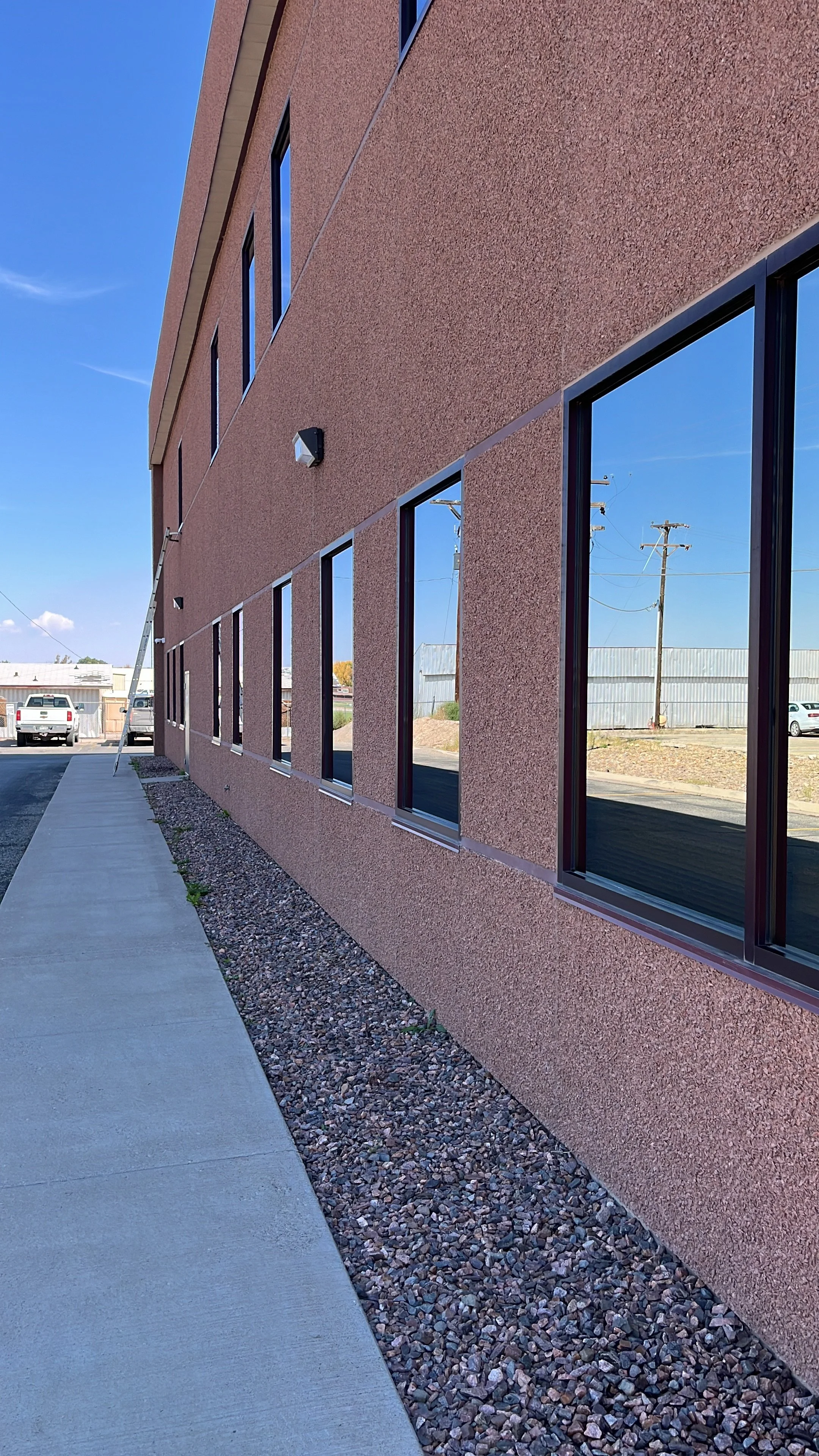 Exterior view of a modern building with pink textured walls and large reflective windows, alongside a sidewalk and gravel strip, with power lines and a parking lot in the background under a blue sky.