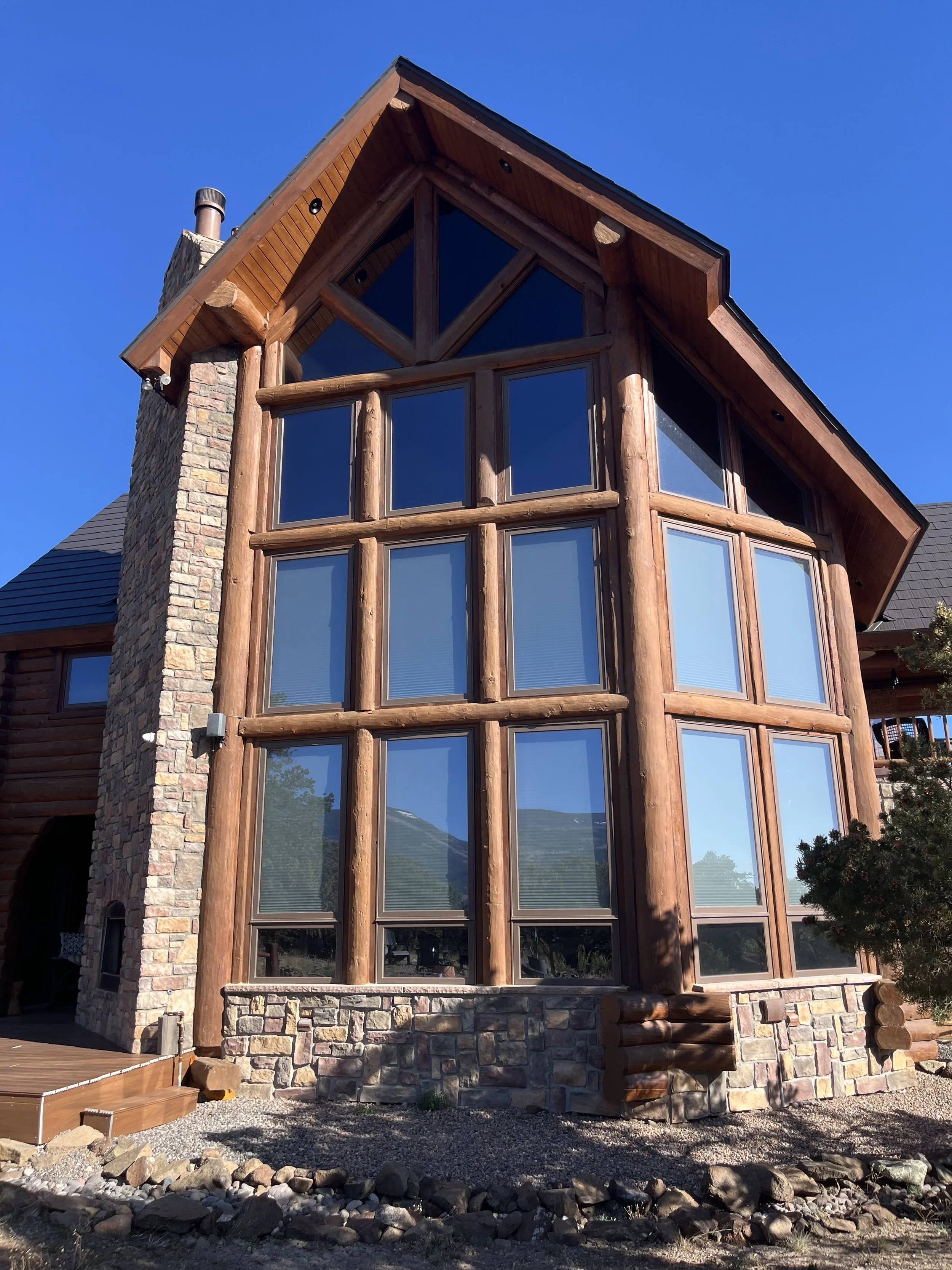 A tall modern house with large windows, a stone chimney, and wooden support beams. The house has a sloped roof, and there is a small set of wooden stairs leading to a deck area. The ground is rocky with some small plants, and a tree is visible on the