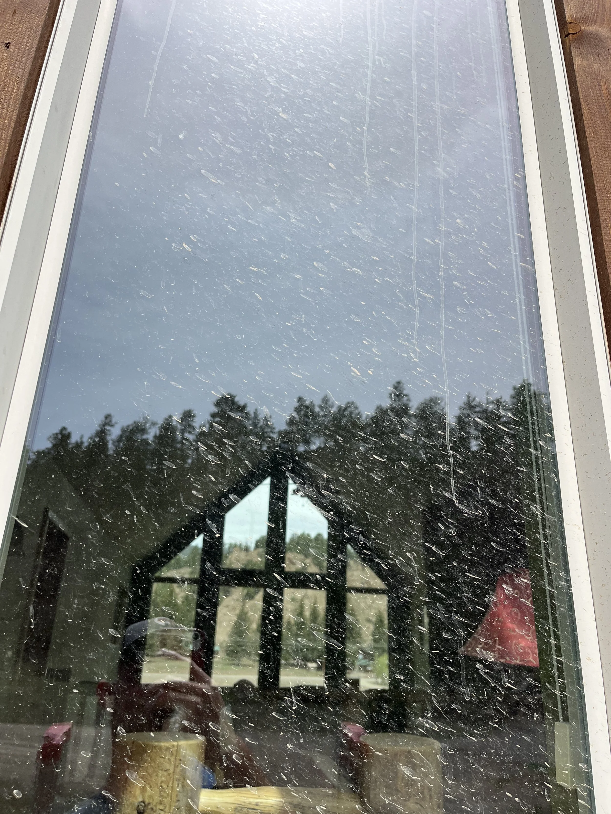 View through a window with raindrops on the glass, showing a cloudy sky and trees outside. The window is framed with white trim, and there are some objects and reflections visible inside.