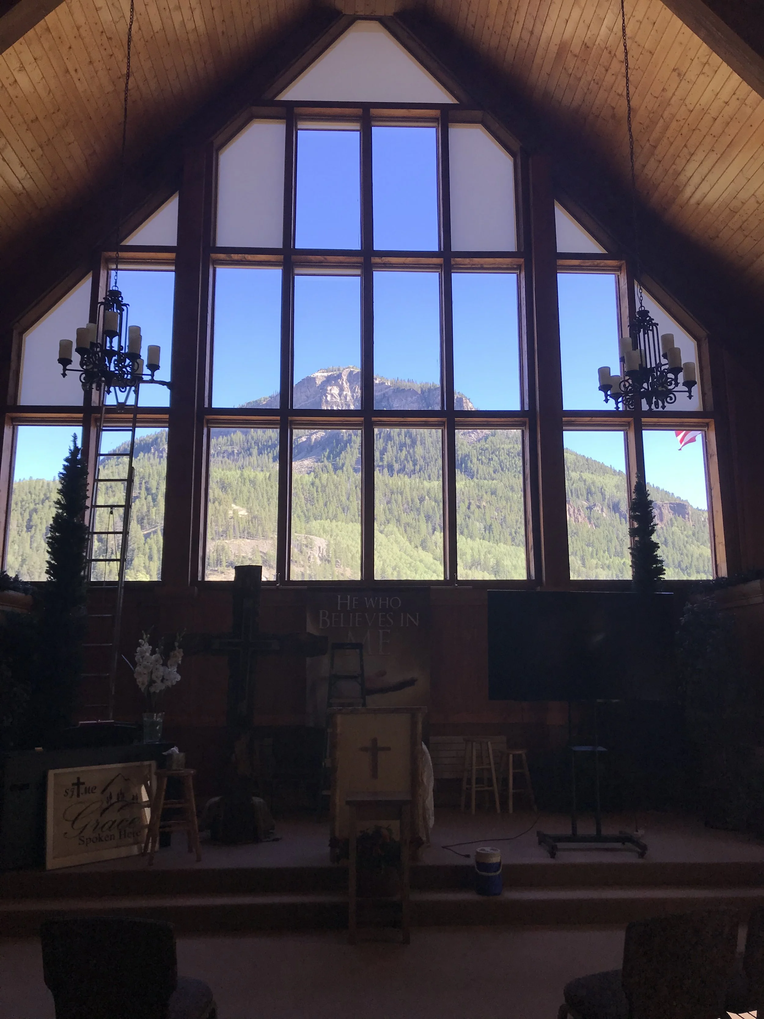 Interior of a church with large wooden-framed stained glass window showing mountain scenery outside. Hanging chandeliers with candles inside. A wooden cross and religious signs are visible.