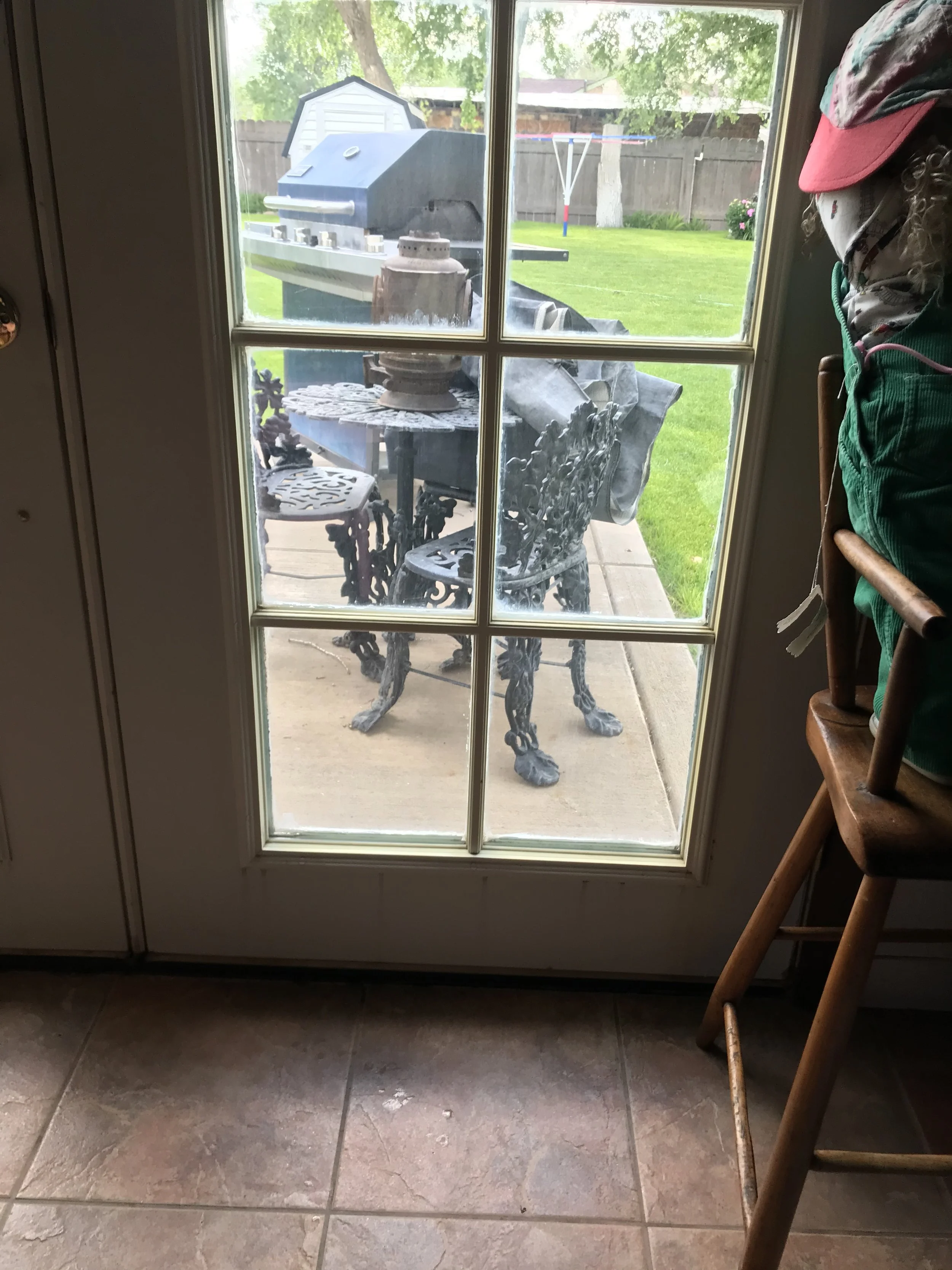 View through a glass door showing a backyard with a barbecue grill, metal outdoor table and chairs, a drying rack, trees, and a wooden fence.