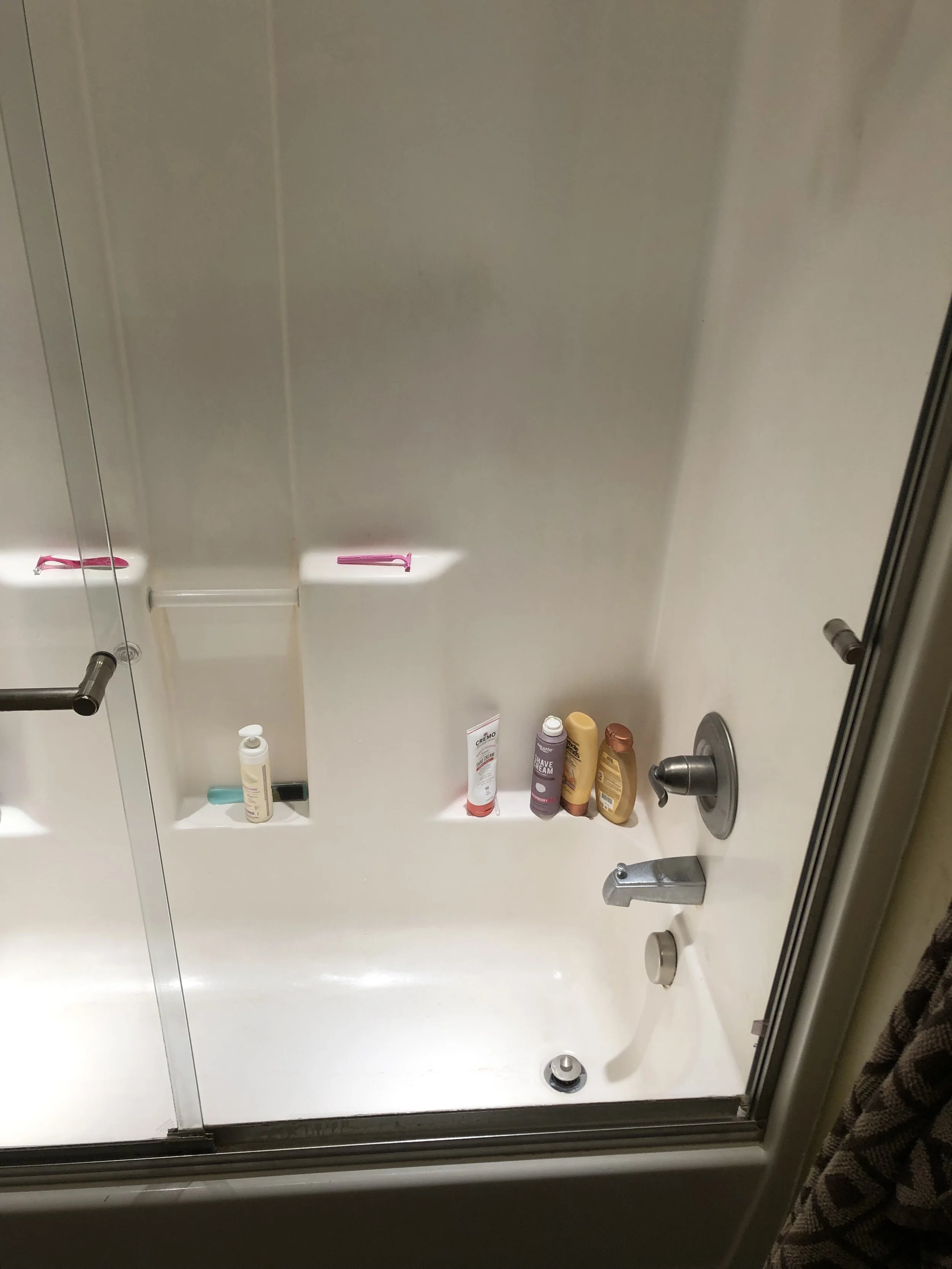 Shower ledge with various toiletries including lotion, shave cream, shampoo, and conditioner bottles, with a showerhead and control knobs visible in a bathroom shower.