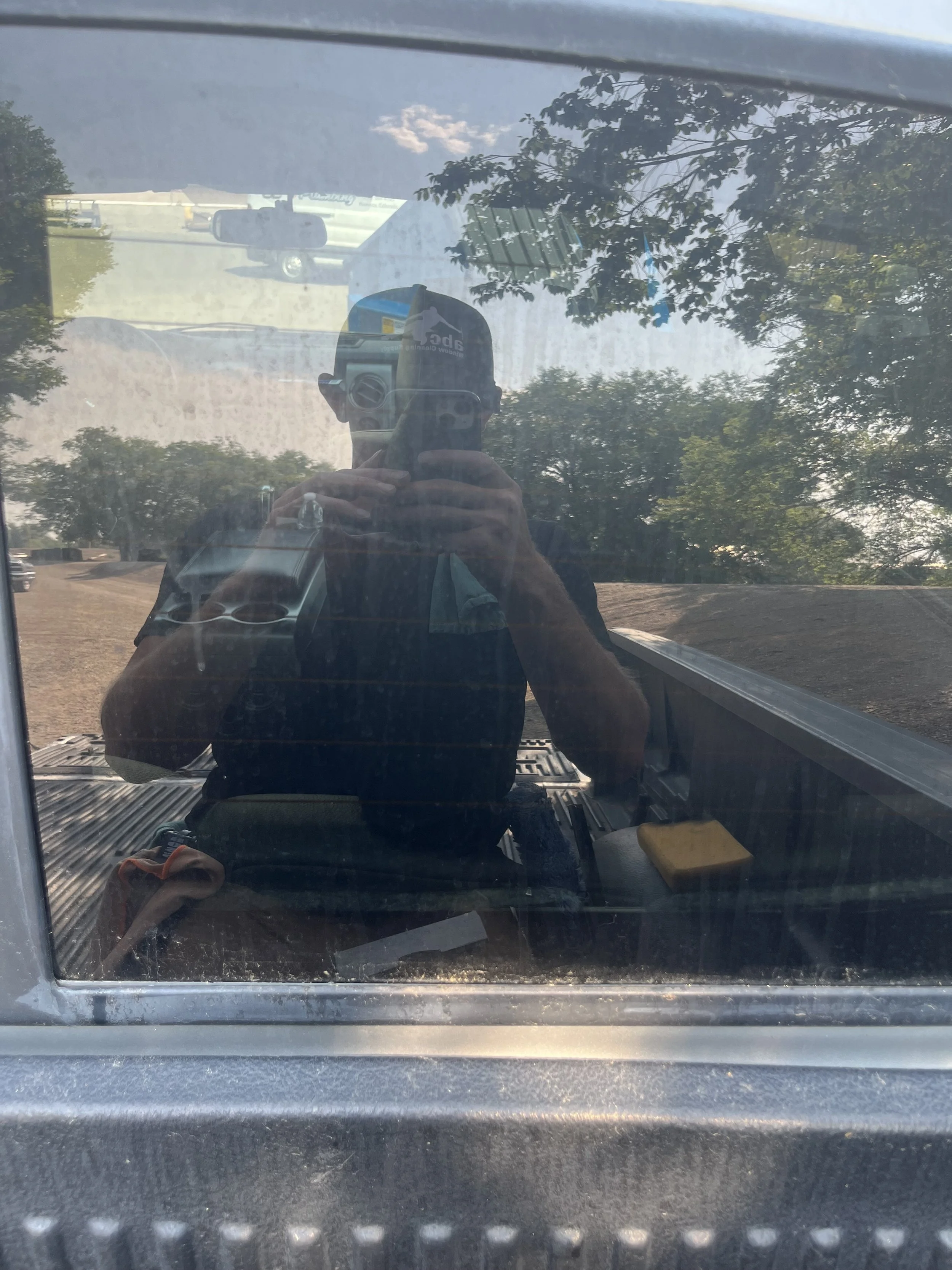 The photo shows a person taking a selfie with a camera in a reflection on a car's hood. There are trees and a parking lot visible in the background.