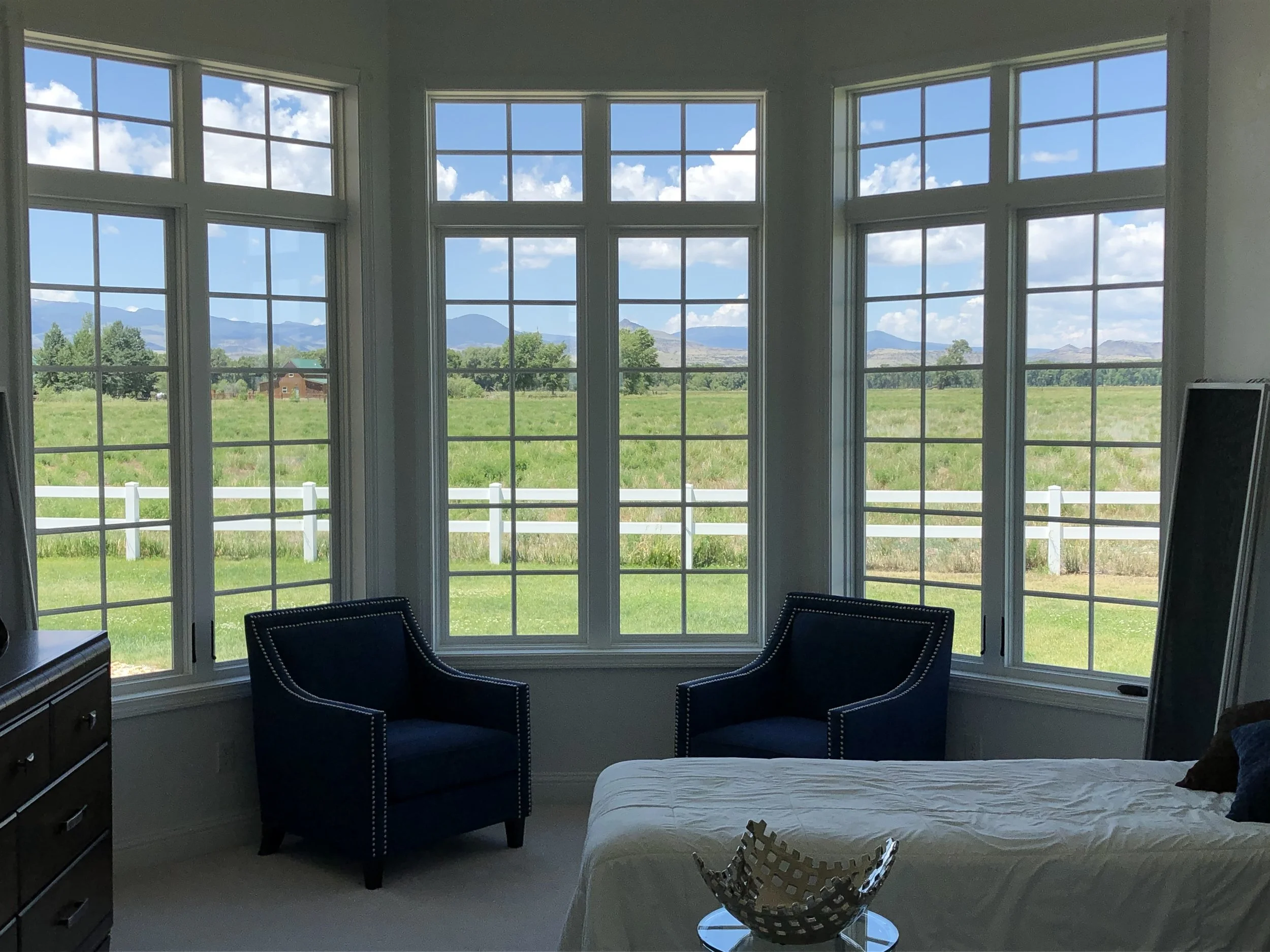 A room with large windows showing a scenic landscape of green fields, trees, and mountains under a blue sky with scattered clouds.
