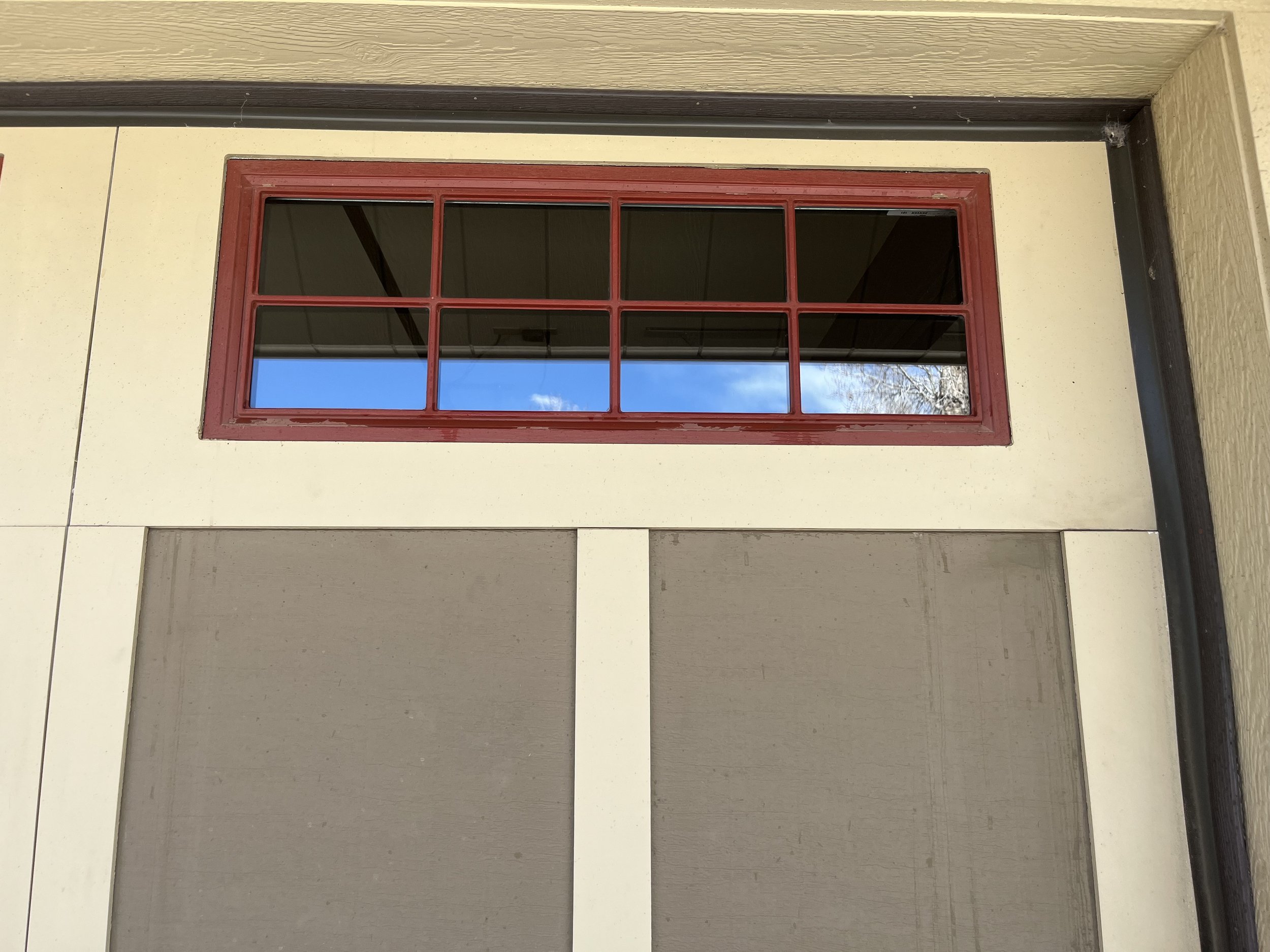 A rectangular window with a red frame installed in a beige wall. The window has eight panes, with the reflection of a clear blue sky and some tree branches visible in the glass. The wall surrounds the window, and there's a darker trim at the top and 