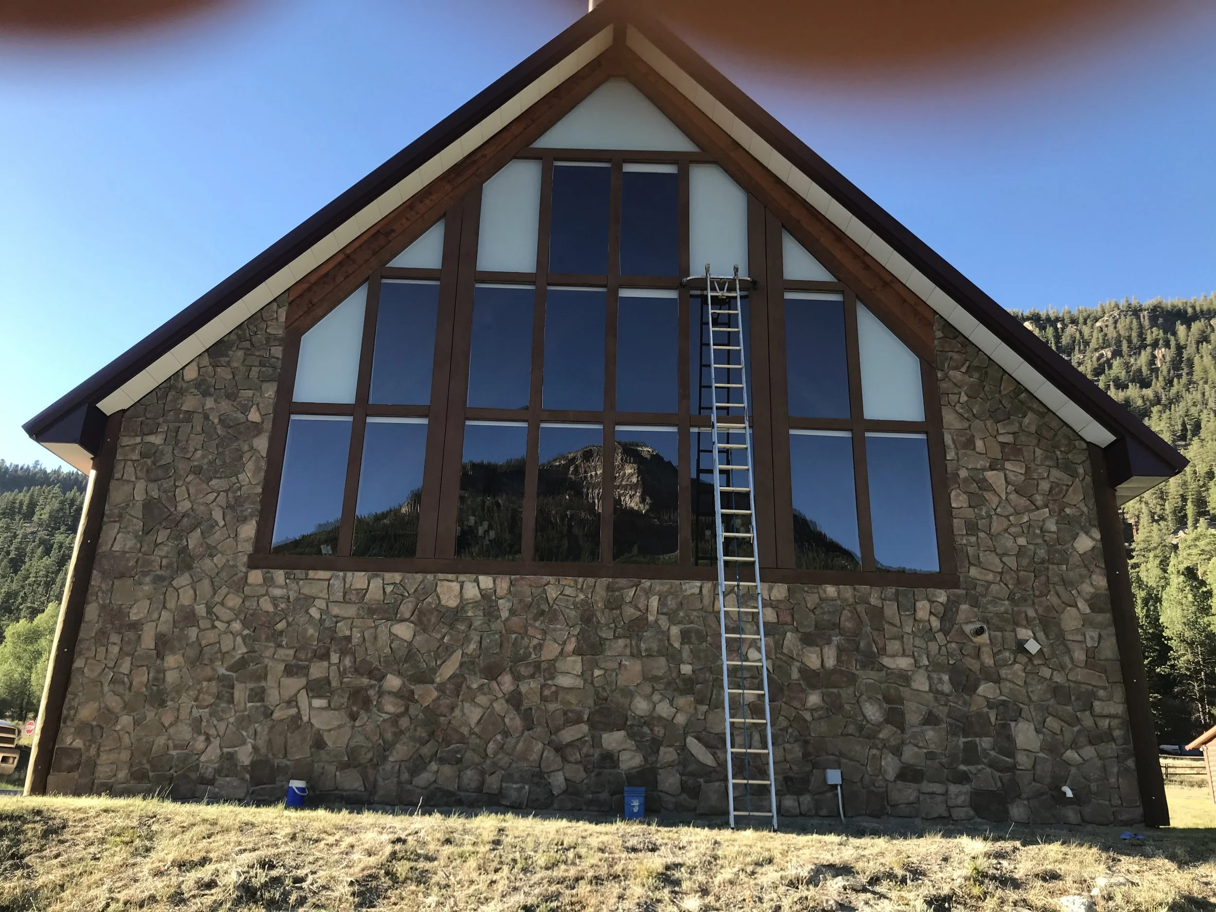 A large house under construction with stone and wood exterior, multiple large windows, and a steep gable roof, with a metal ladder leaning against it and a clear blue sky in the background.