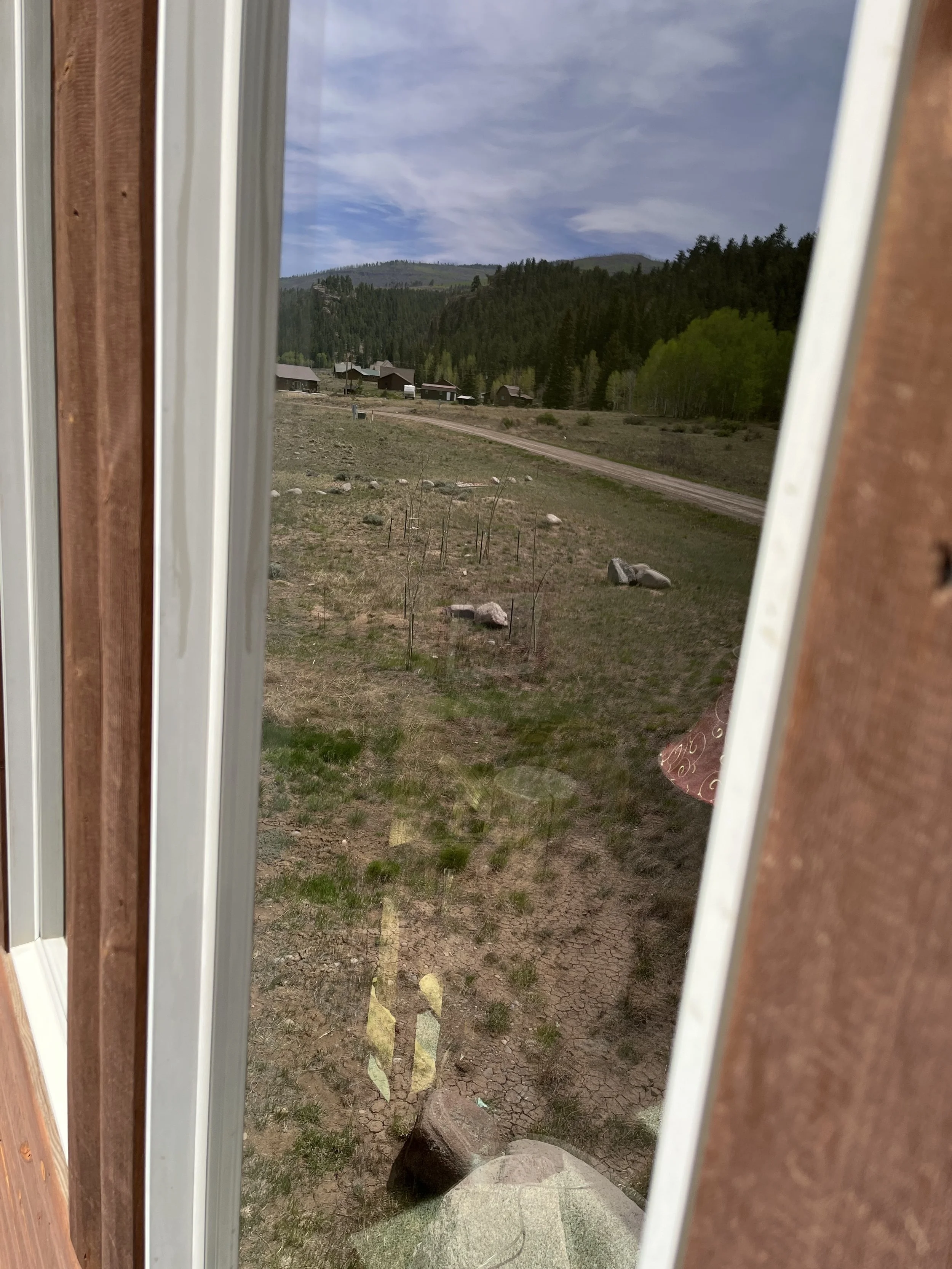 View of a rural landscape with a dirt road, scattered rocks, and small houses surrounded by green trees and mountains in the background, seen through a window.