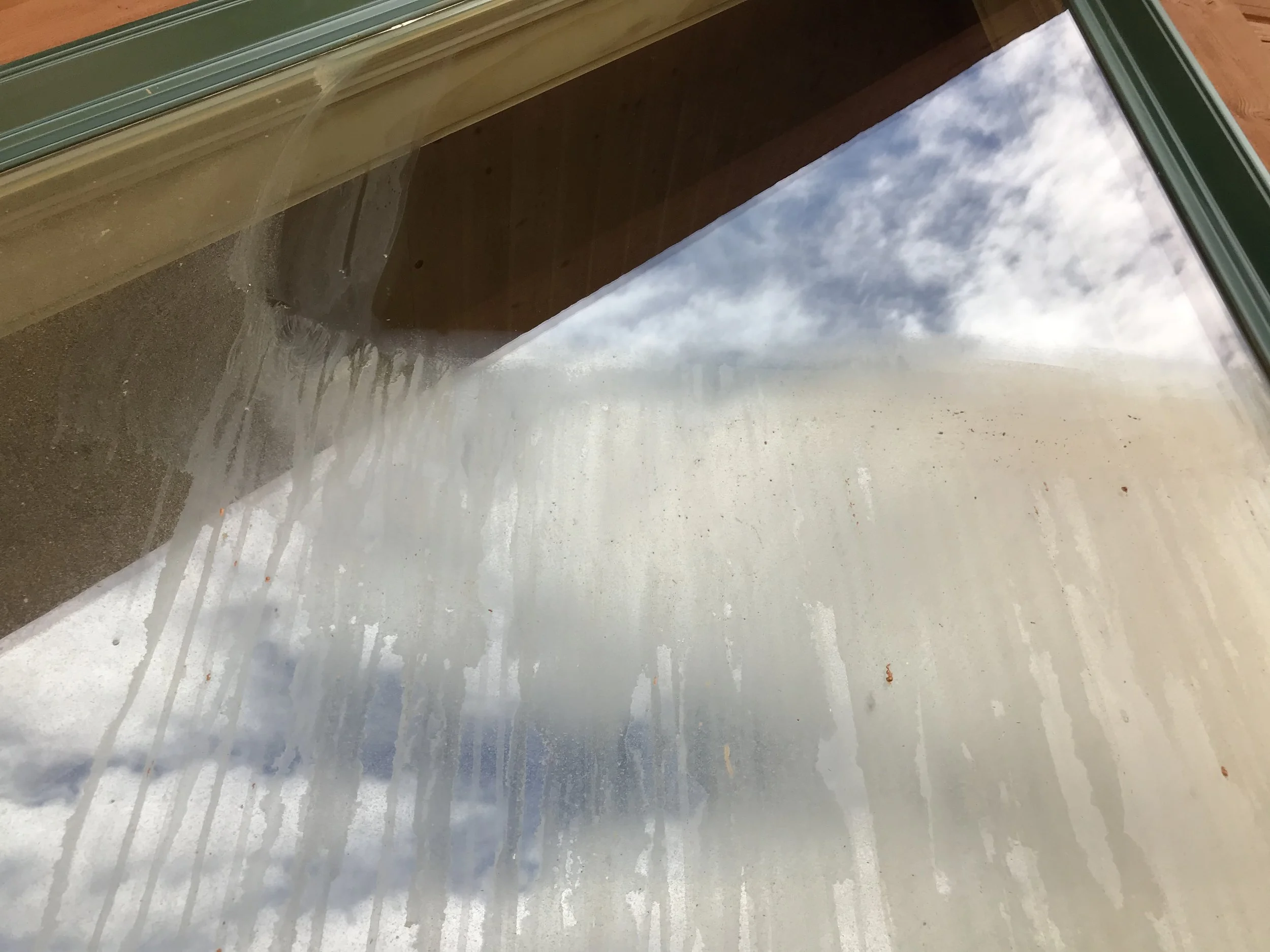 Looking through a glass window or door with a view of a cloudy sky outside. The interior part of the window or door has a green frame and some dirt or water spots.